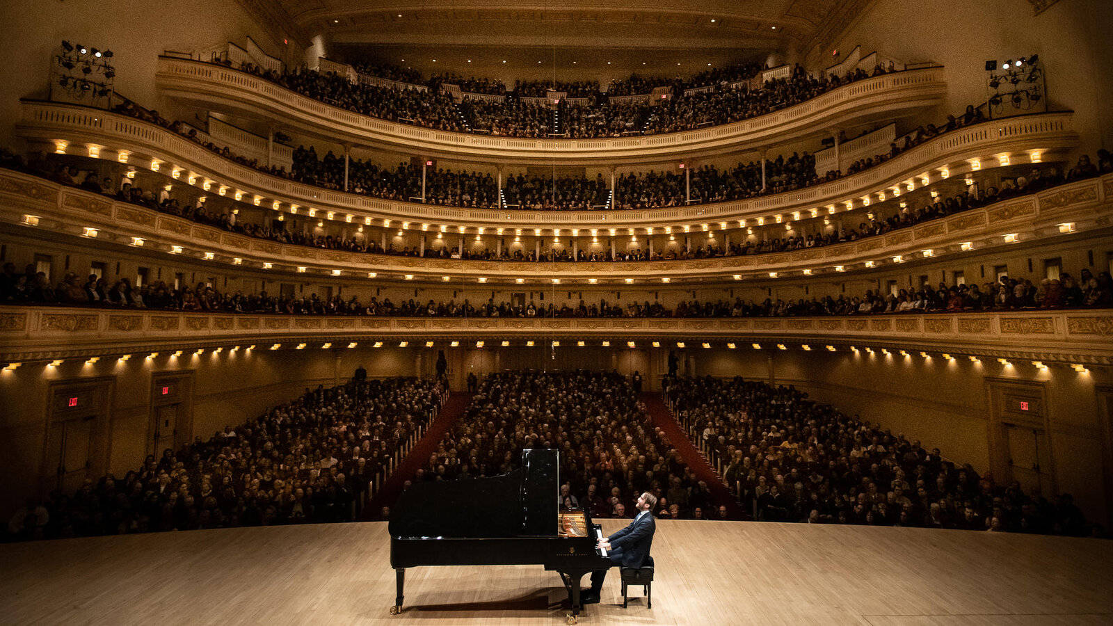 concert 桌布 壁紙 — 一位孤獨的鋼琴家在寬敞華麗的音樂廳中，在溫暖的金色燈光下，於分層座無虛席的觀眾前演奏三角鋼琴。
