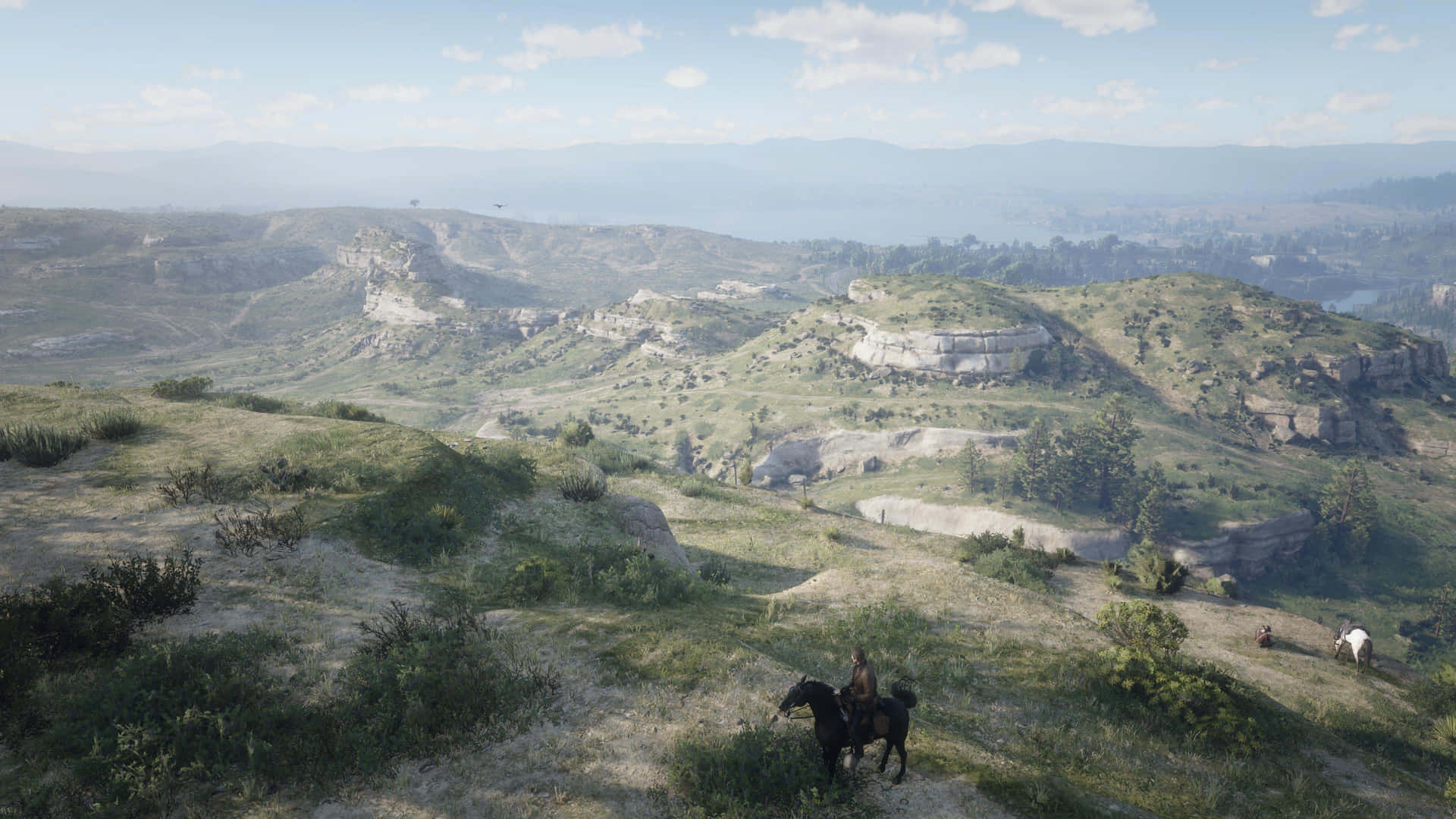 Red Dead Redemption 4K 桌布 壁紙 — 一位孤獨的騎手騎著一匹黑馬，穿越陽光明媚的草地山坡，俯瞰廣闊的起伏丘陵和遠處的山脈，天空部分多雲。