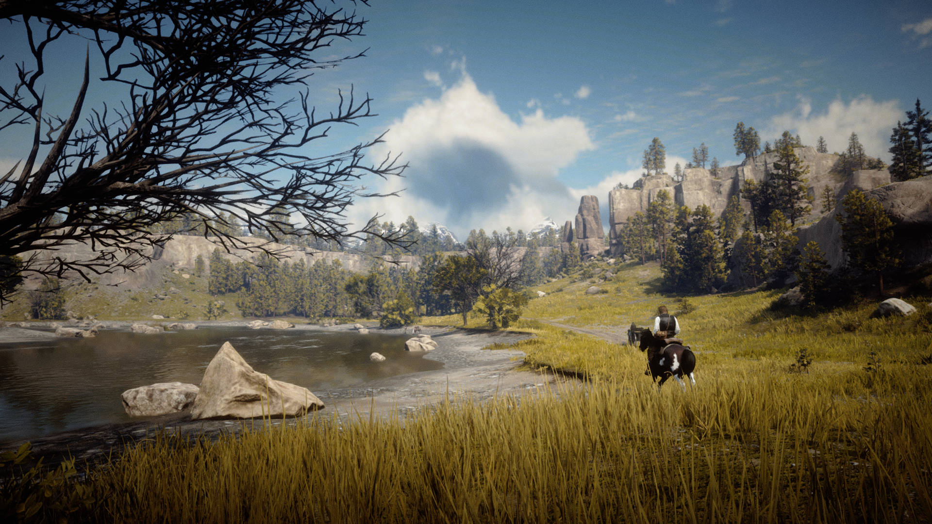 Red Dead Redemtion 2 桌布 壁紙 — 一位孤獨的騎手騎著馬，在明亮的藍天下，穿過長滿草的河岸，天空中有散落的雲朵和遠處的岩石峭壁。