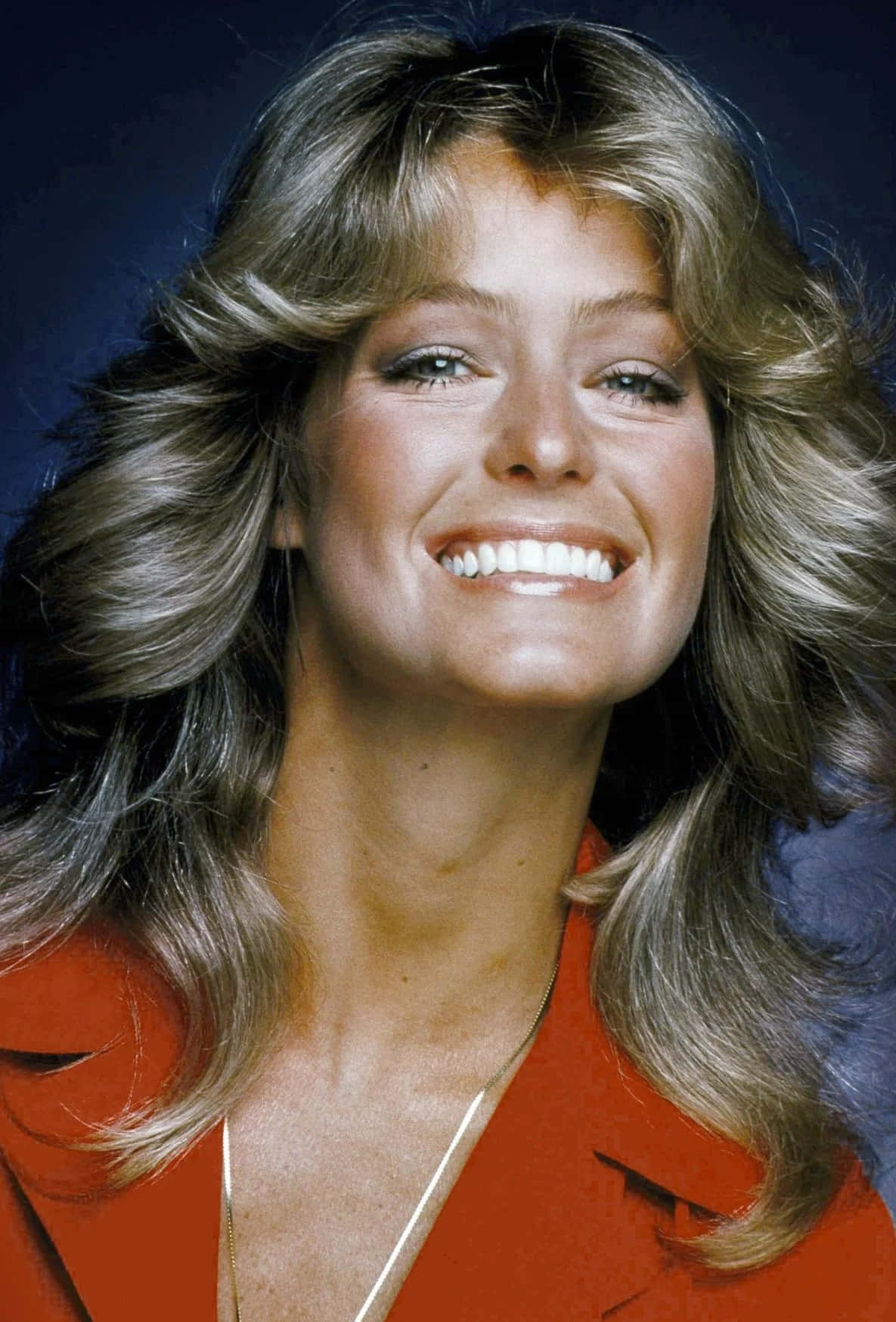 Farrah Fawcett 桌布 壁紙 — 一位容光煥發、擁有一頭豐盈金髮的女性，在深藍色攝影棚背景下穿著大膽的紅色外套燦爛微笑。