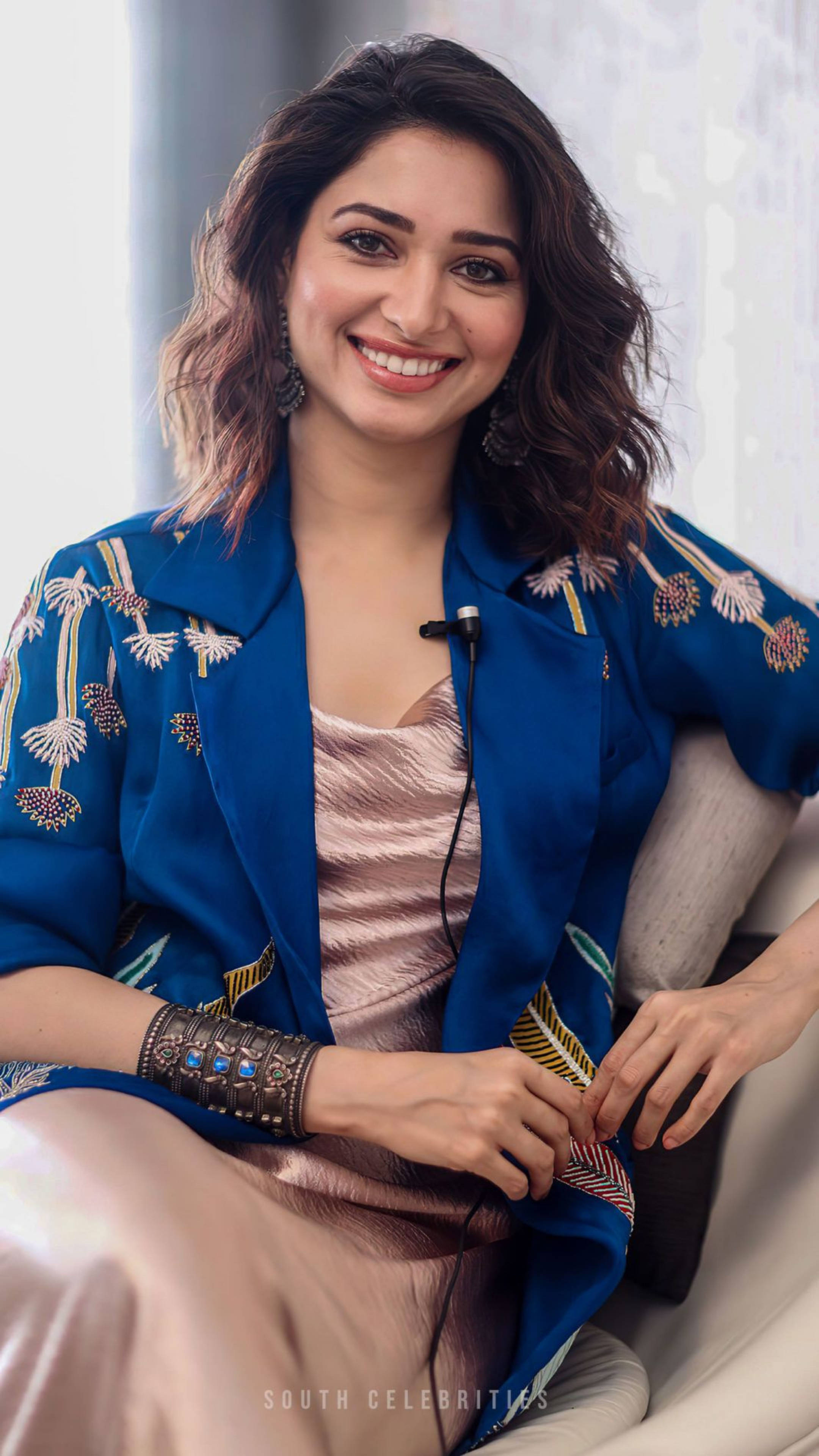 tamannaah bhatia 桌布 壁紙 — 一位容光煥發、擁有波浪棕色頭髮的女性，穿著藍色刺繡外套和金屬感上衣，坐在椅子上溫暖地微笑。