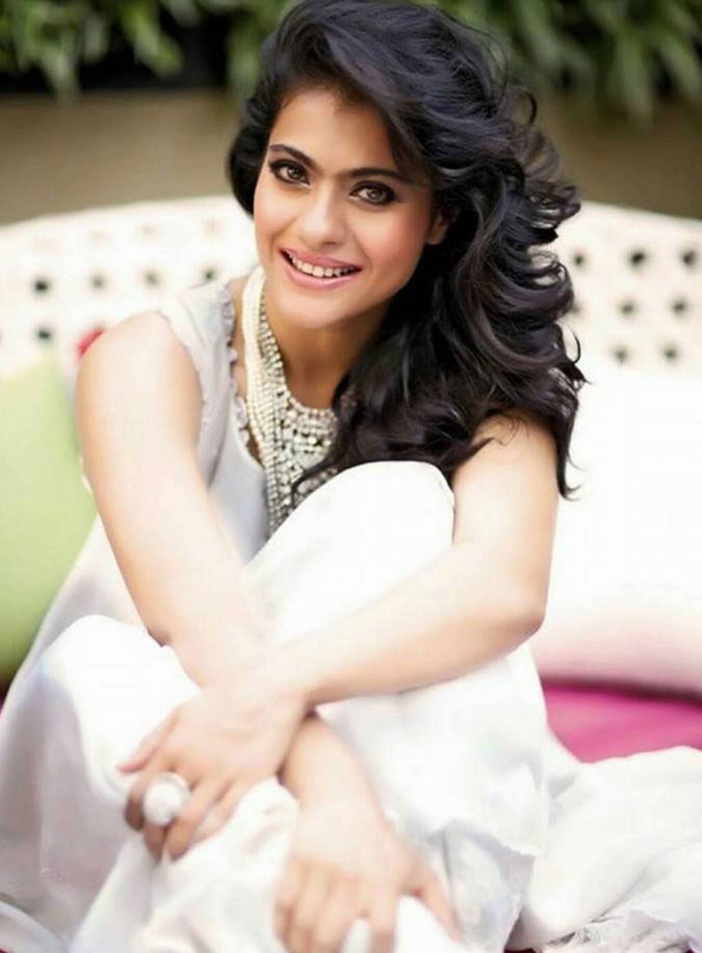 Kajol 桌布 壁紙 — 一位容光煥發、擁有深色波浪長髮的女性，身穿白色服裝和分層首飾，坐在有圖案的戶外沙發上，溫暖地微笑。