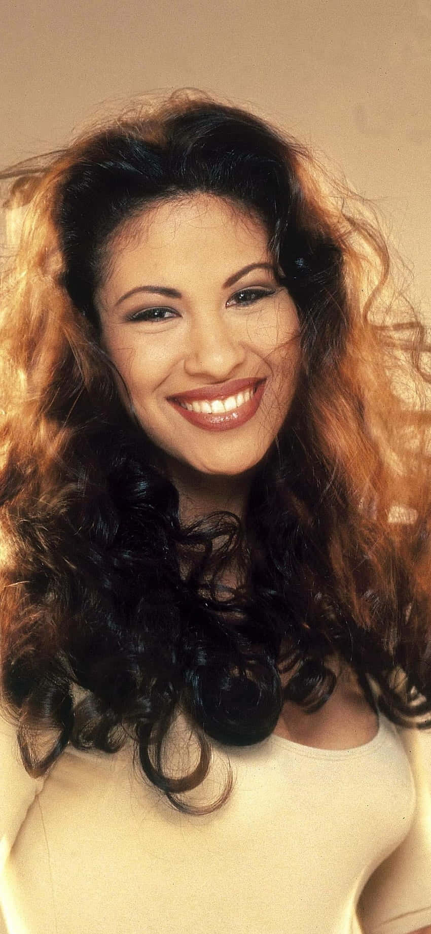 Selena Quintanilla iPhone 桌布 壁紙 — 一位容光煥發、擁有豐盈捲髮的女性在柔和的米色背景前溫暖地微笑。