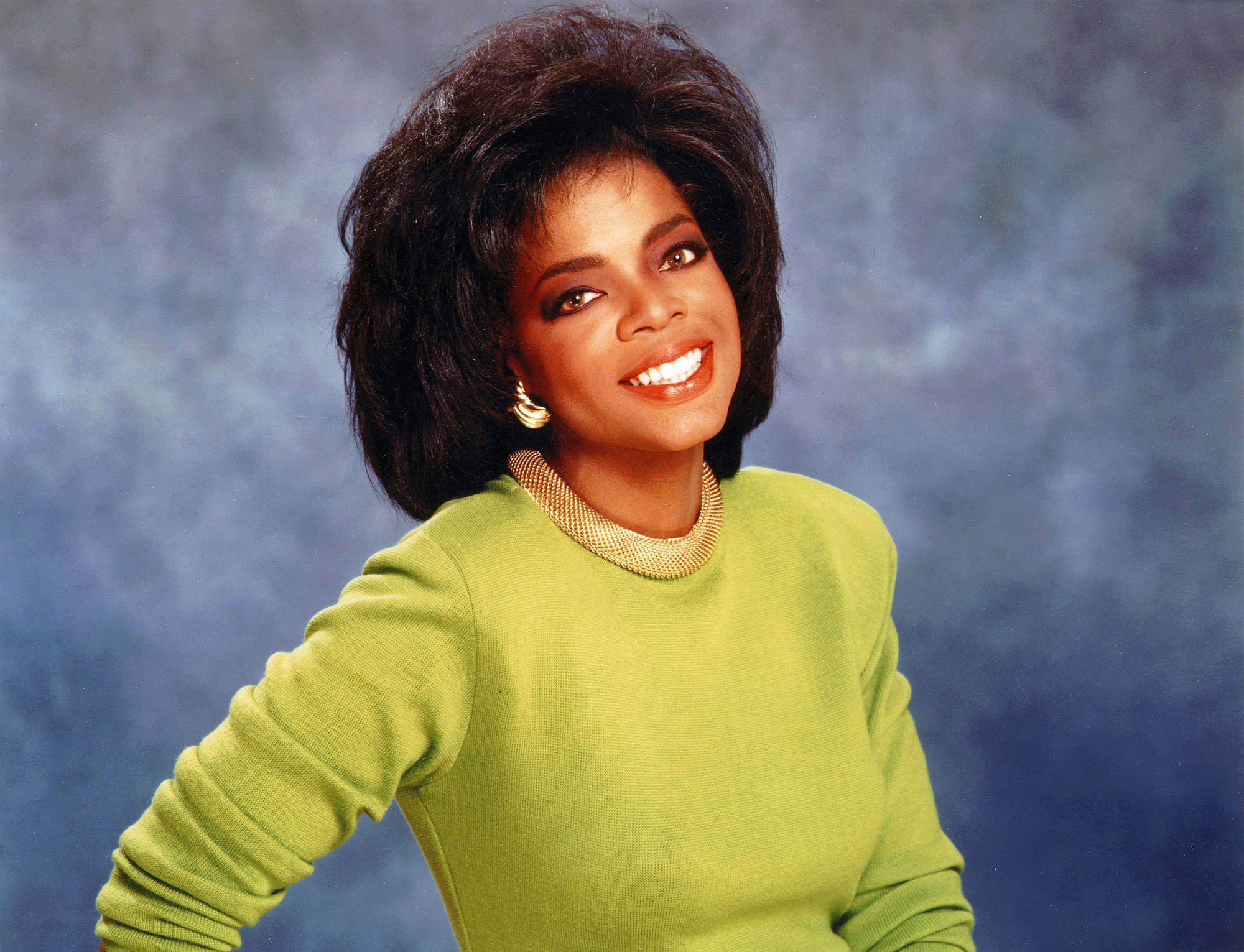 Oprah 桌布 壁紙 — 一位容光煥發、擁有豐盈深色頭髮的女性，穿著亮綠色毛衣，在帶有紋理的藍色背景前溫暖地微笑。