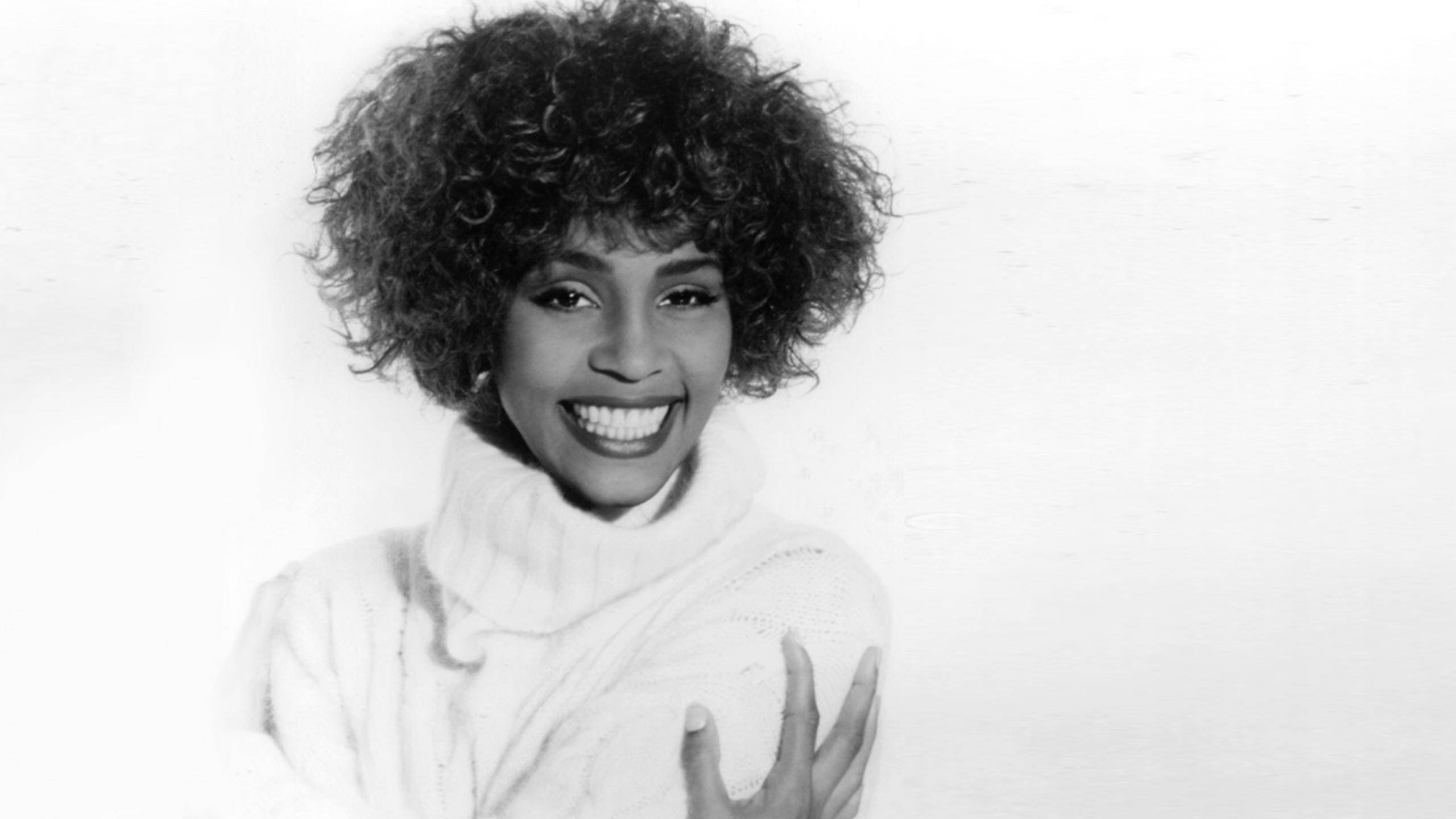 Whitney 桌布 壁紙 — 一位容光煥發、擁有豐盈爆炸頭的女性，在純色背景前穿著白色高領毛衣，燦爛地微笑。