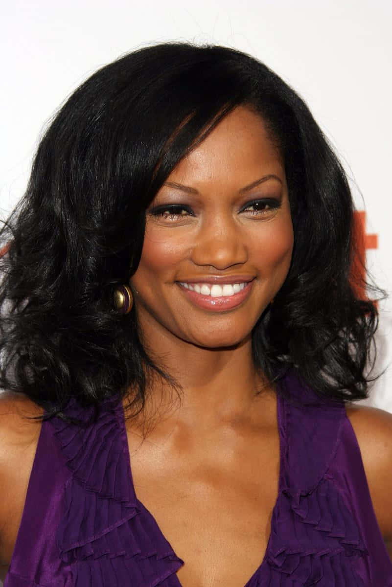 Garcelle Beauvais 桌布 壁紙 — 一位容光煥發、擁有豐盈黑髮和燦爛笑容的女性，在純白背景前穿著荷葉邊紫色上衣。