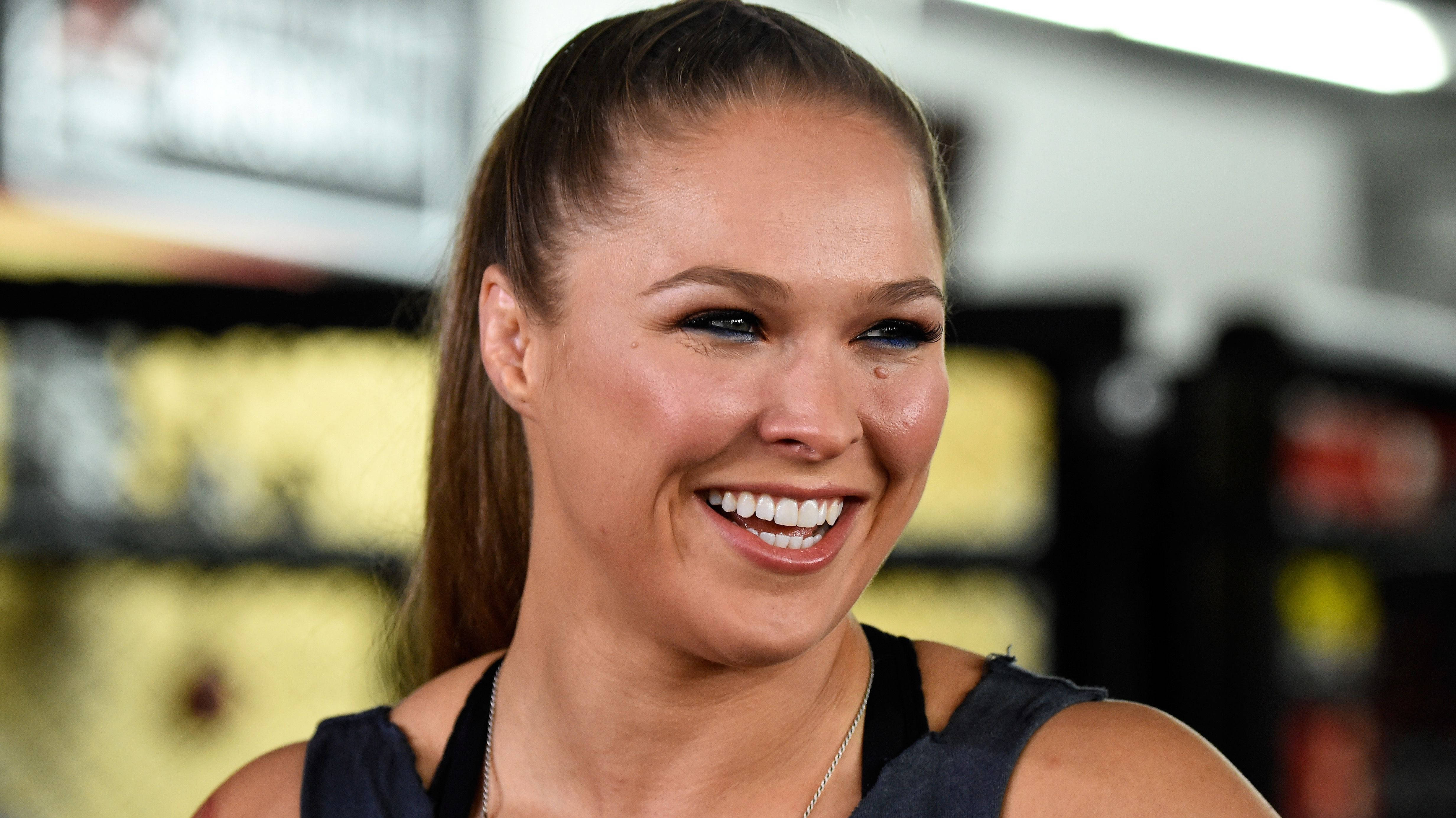 Rousey 桌布 壁紙 — 一位容光煥發、笑容燦爛、紮著馬尾辮的女性在健身房環境中看向鏡頭外。
