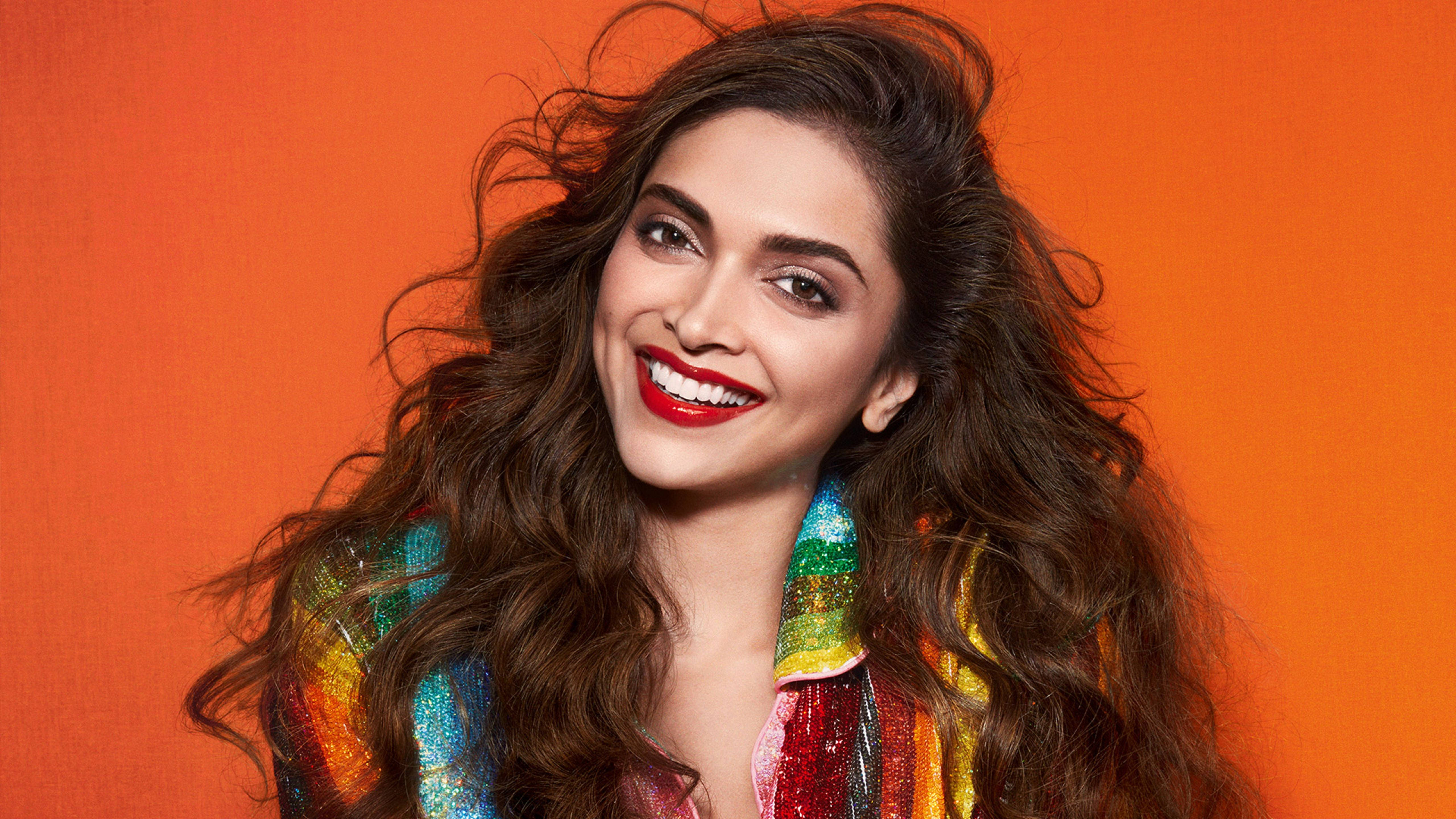 Deepika 桌布 壁紙 — 一位容光煥發的女性，擁有波浪般的棕色頭髮和鮮豔的紅色唇膏，在充滿活力的橙色背景前微笑，穿著色彩繽紛的亮片上衣。