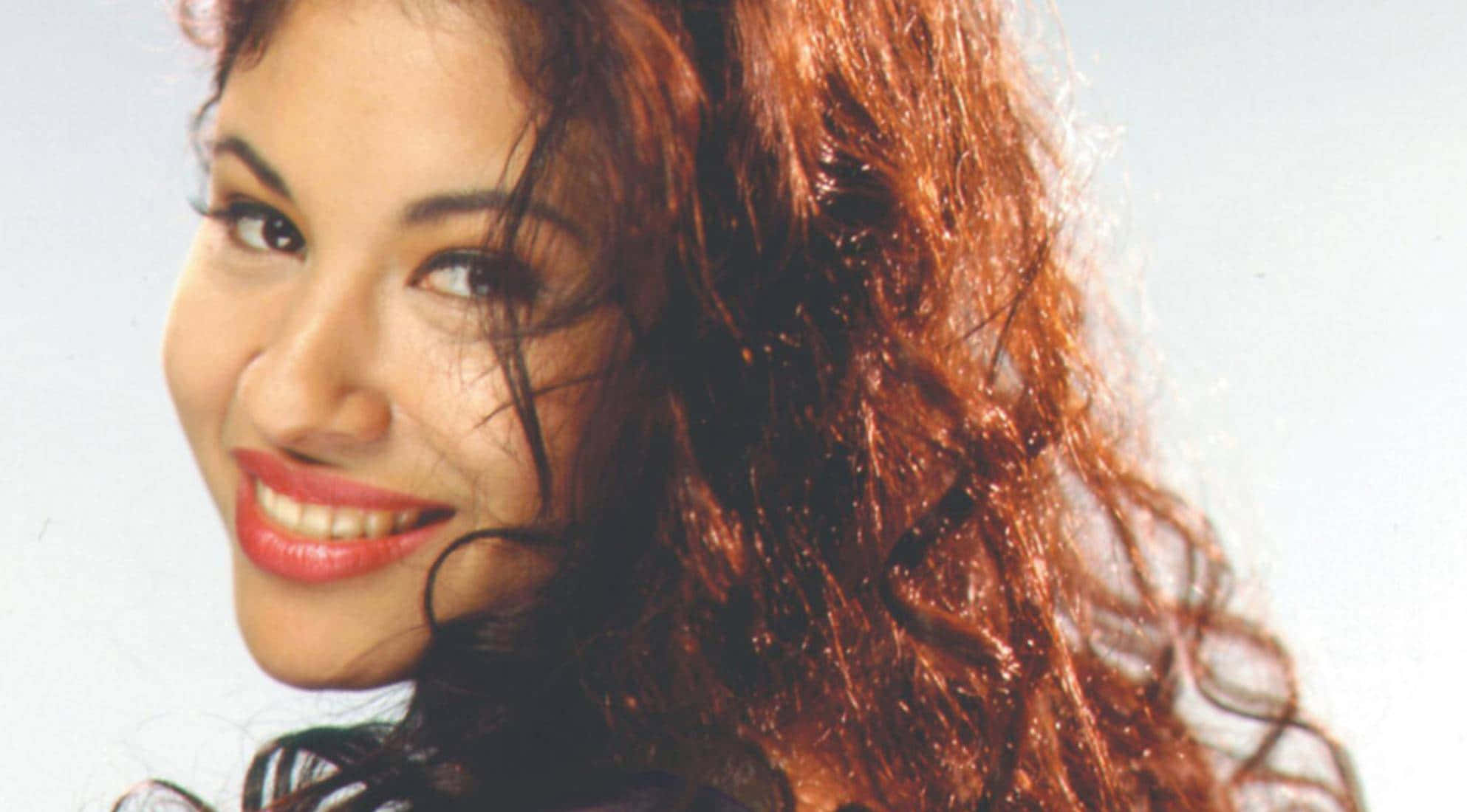 Selena Quintanilla iPhone 桌布 壁紙 — 一位容光煥發的女性，擁有豐盈的赤褐色捲髮，在柔和的白色背景前溫暖地微笑。