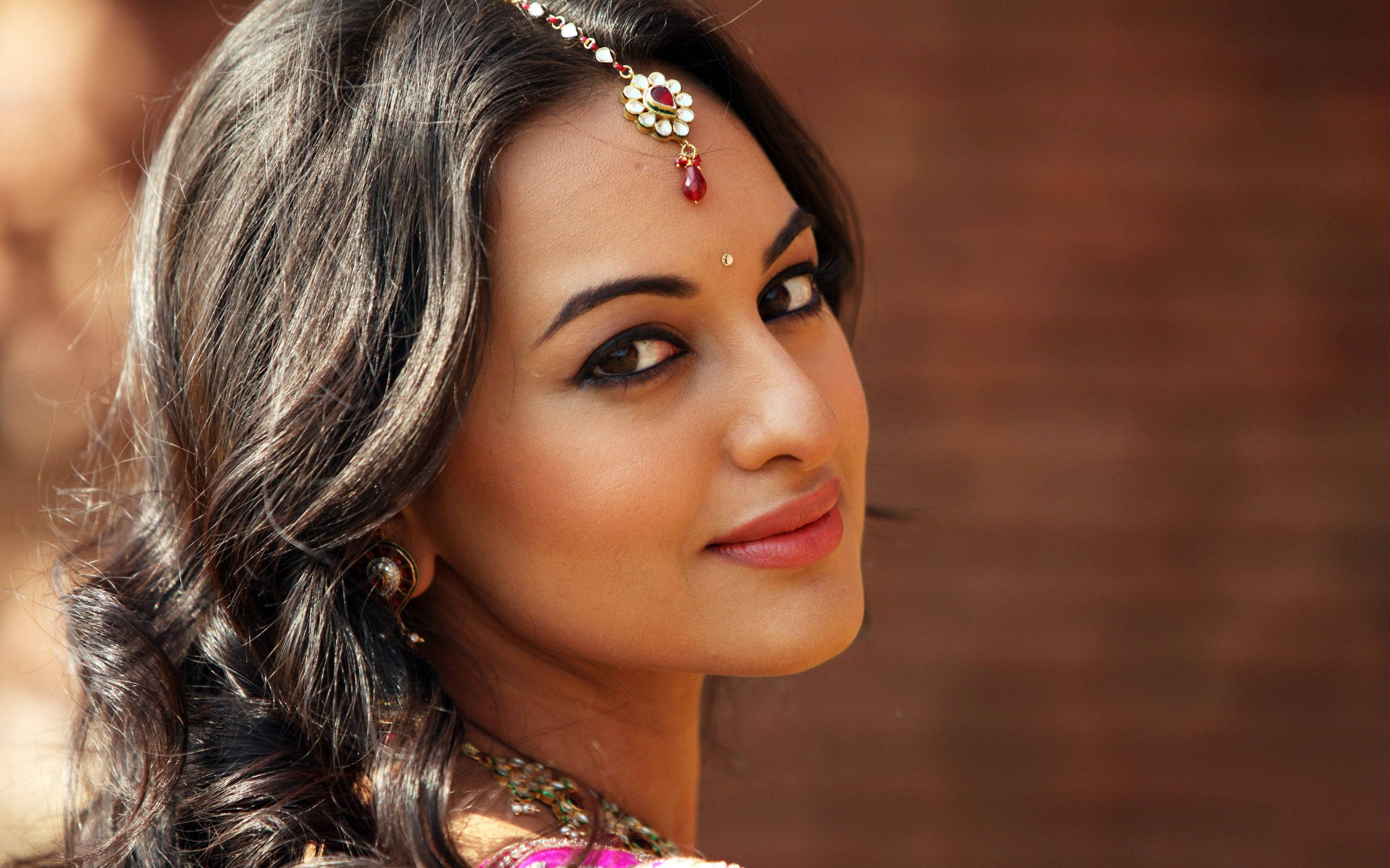 Sonakshi Sinha 桌布 壁紙 — 一位容光煥發的女性，深色波浪髮上飾有紅色寶石Maang Tikka，在溫暖模糊的磚牆背景下溫柔微笑。