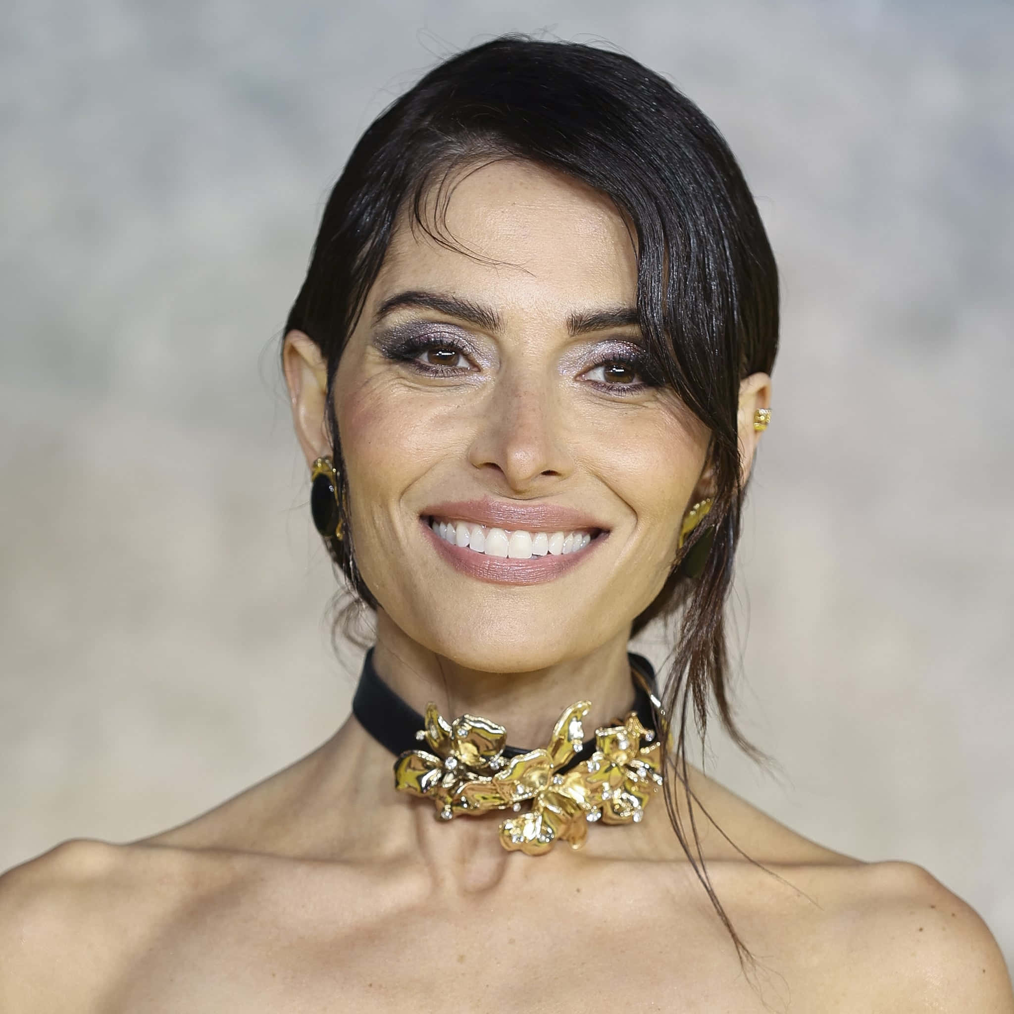 sarah shahi 桌布 壁紙 — 一位容光煥發的女性在柔和的灰色背景下溫暖微笑，佩戴著大膽的金色花卉頸鏈和相配的耳環。