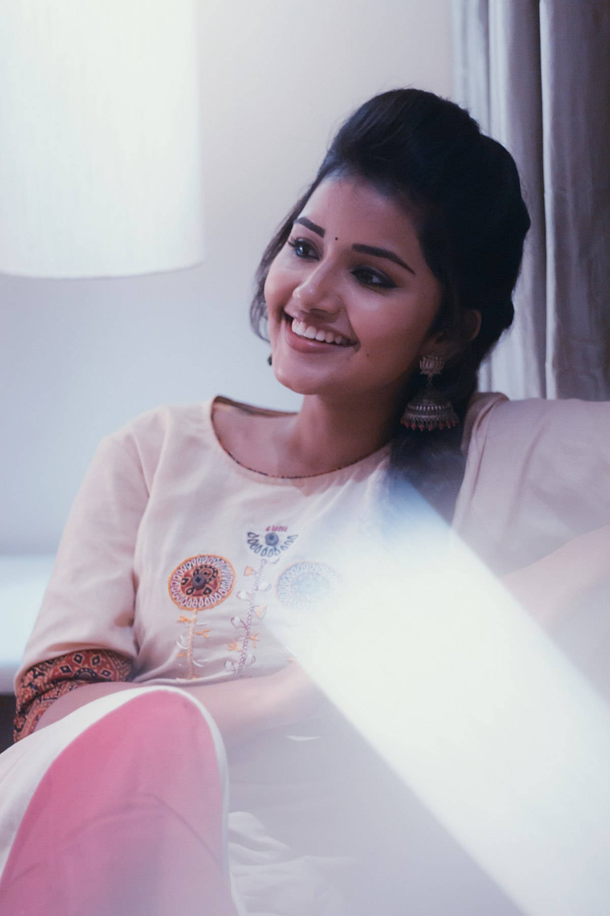 anupama parameswaran 桌布 壁紙 — 一位容光煥發的女性坐在室內，身旁窗戶透進陽光，她溫暖地微笑。