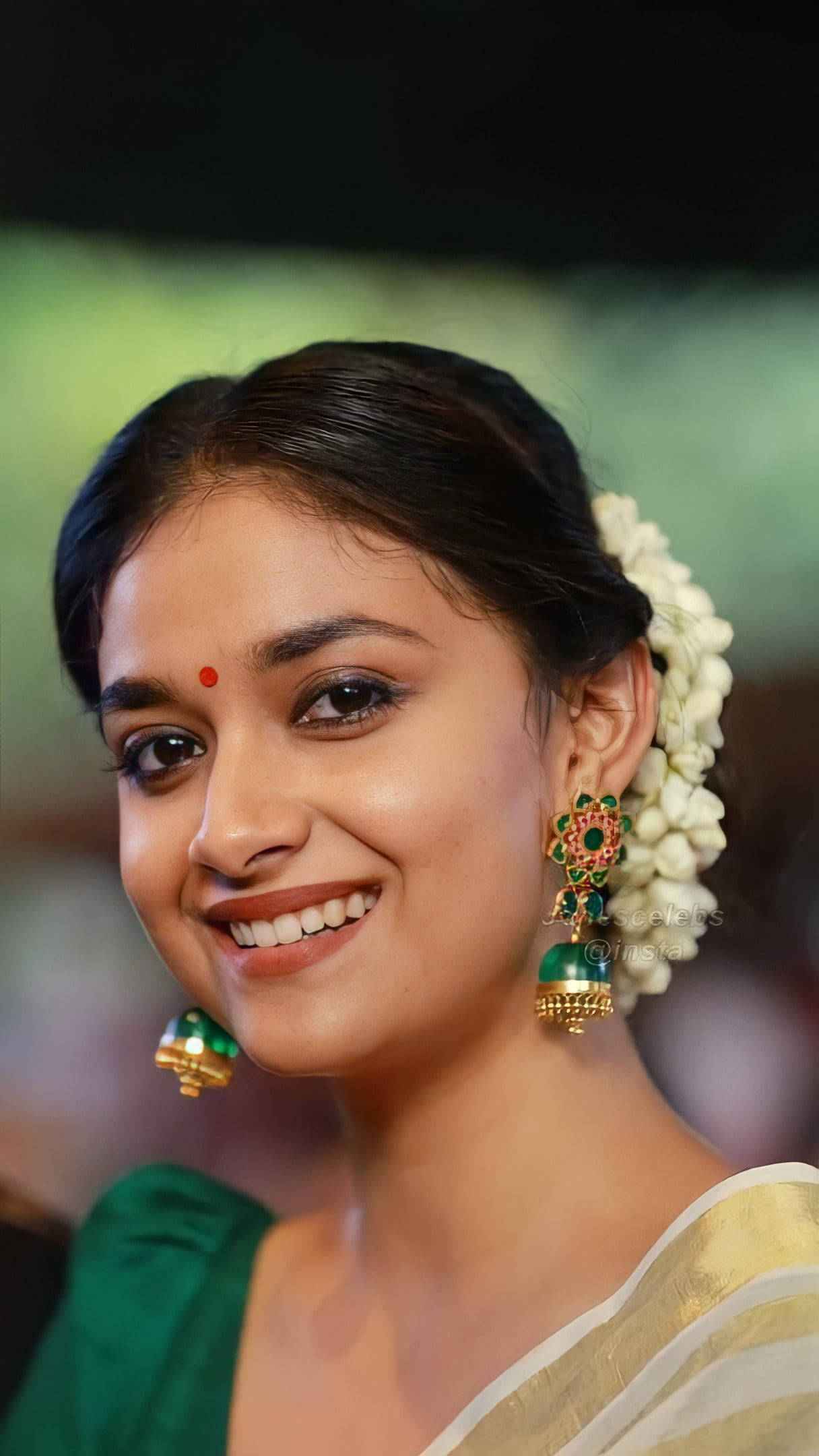 telugu 桌布 壁紙 — 一位容光煥發的女性溫暖微笑，頭戴花卉髮飾，佩戴華麗綠色耳環，背景柔和模糊。