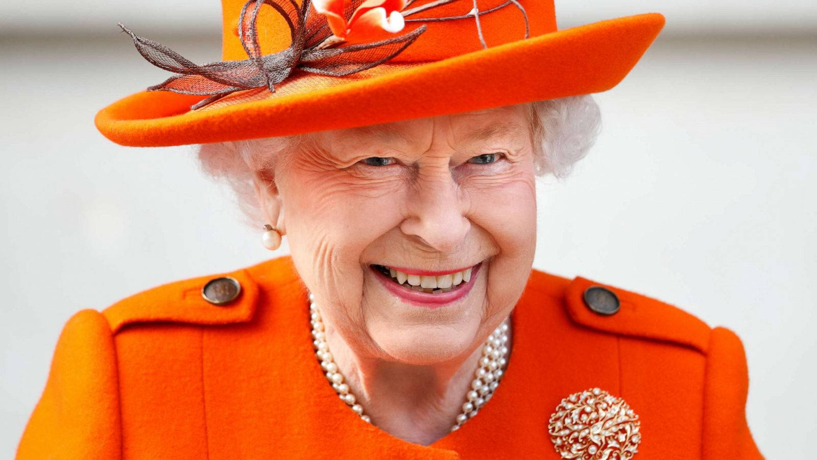 Queen Elizabeth 桌布 壁紙 — 一位容光煥發的老年女性身穿鮮豔的橙色外套和相配的帽子，帽子上裝飾著花卉點綴，笑容滿面。