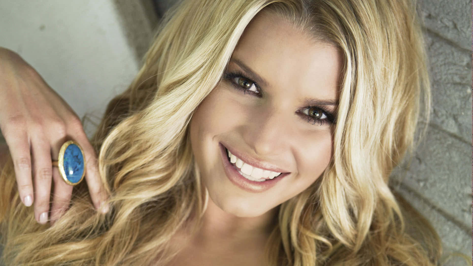 jessica simpson 桌布 壁紙 — 一位容光煥發的金髮女子，面帶燦爛笑容，手上戴著一個大藍色戒指，在紋理石牆前擺姿勢。