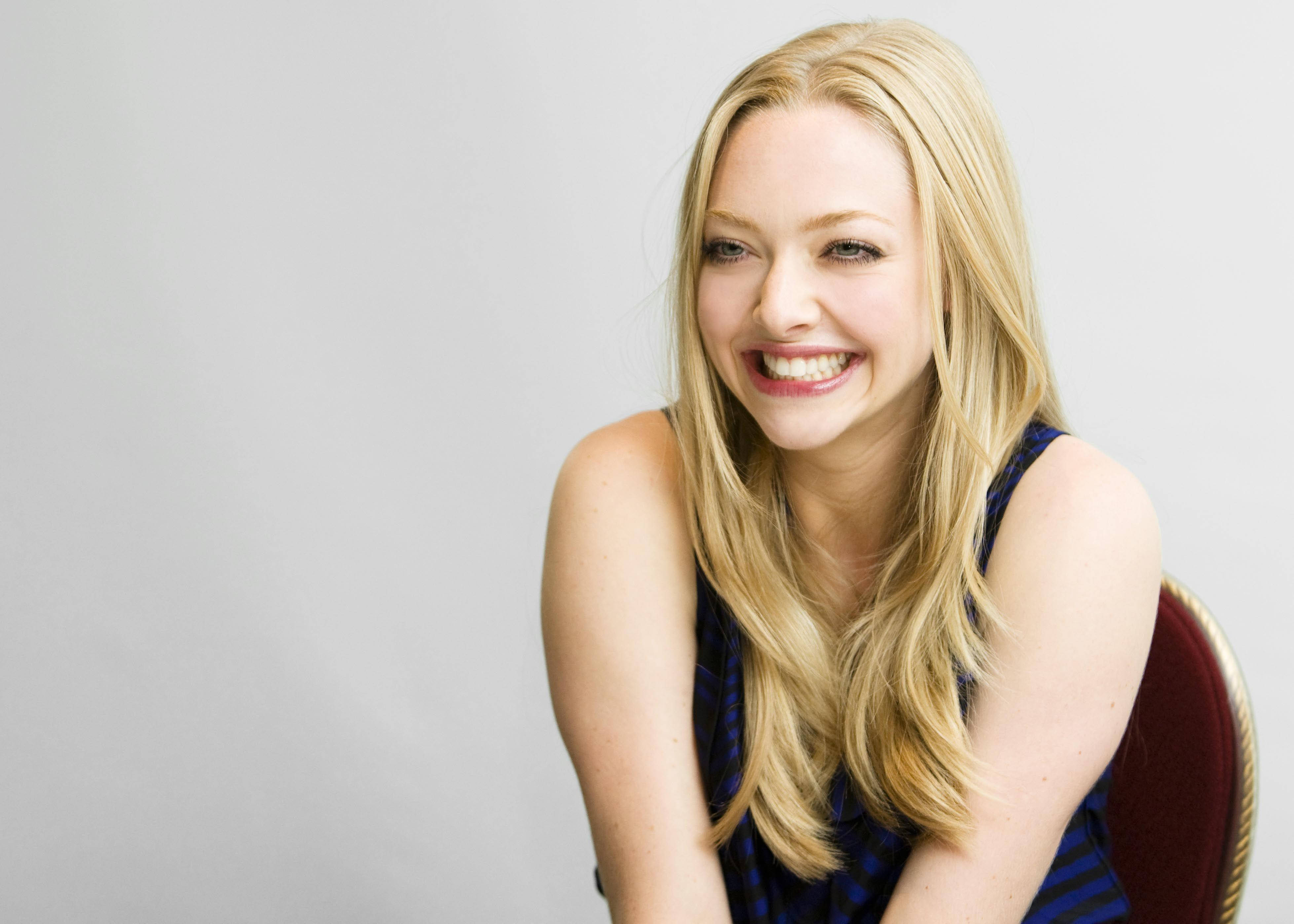 Amanda Seyfried 桌布 壁紙 — 一位容光煥發的金髮女子面帶燦爛笑容，坐在紅色椅子上，背景是純灰色。