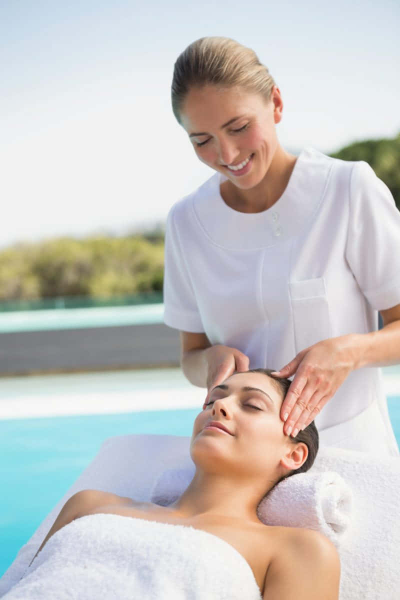Massage 桌布 壁紙 — 一位寧靜的女性在明亮的日光下，於閃爍的泳池旁接受微笑治療師的放鬆面部按摩。