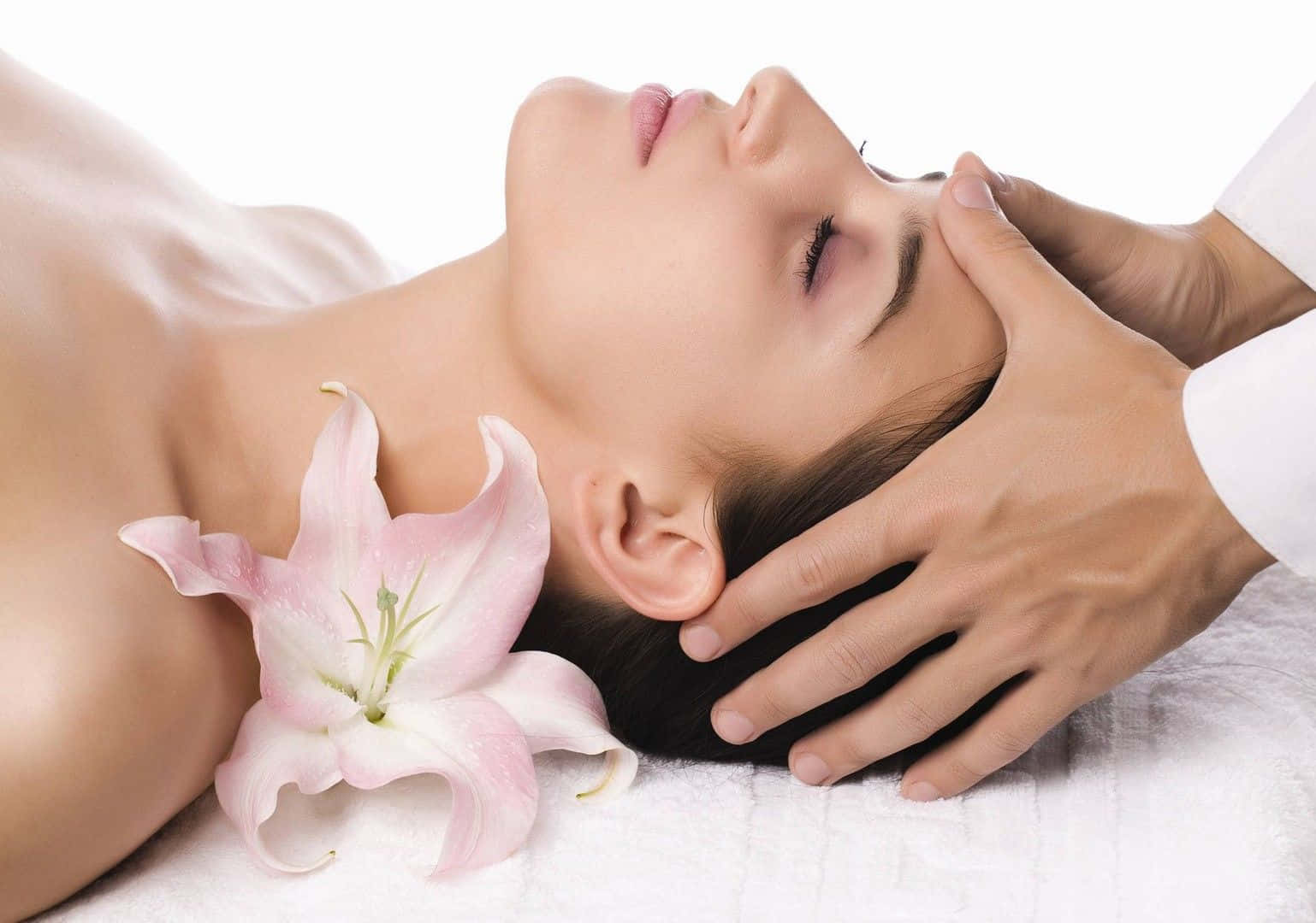 Massage 桌布 壁紙 — 一位寧靜的女性正在接受輕柔的頭部按摩，胸前放著一朵粉紅色的百合花，背景是純白色。