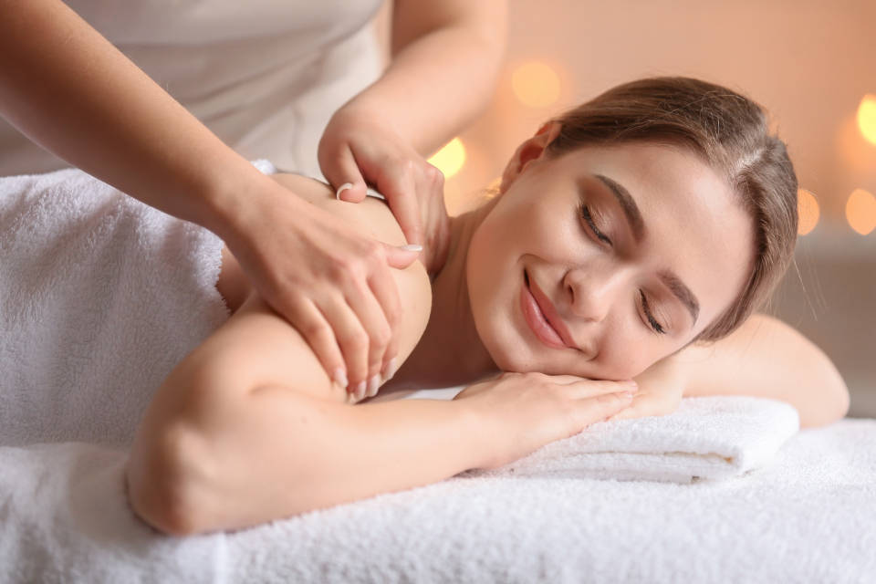 Relaxing 桌布 壁紙 — 一位寧靜的女性閉著眼睛微笑，治療師在柔和燈光及溫暖散景燈光的SPA中按摩她的肩膀。