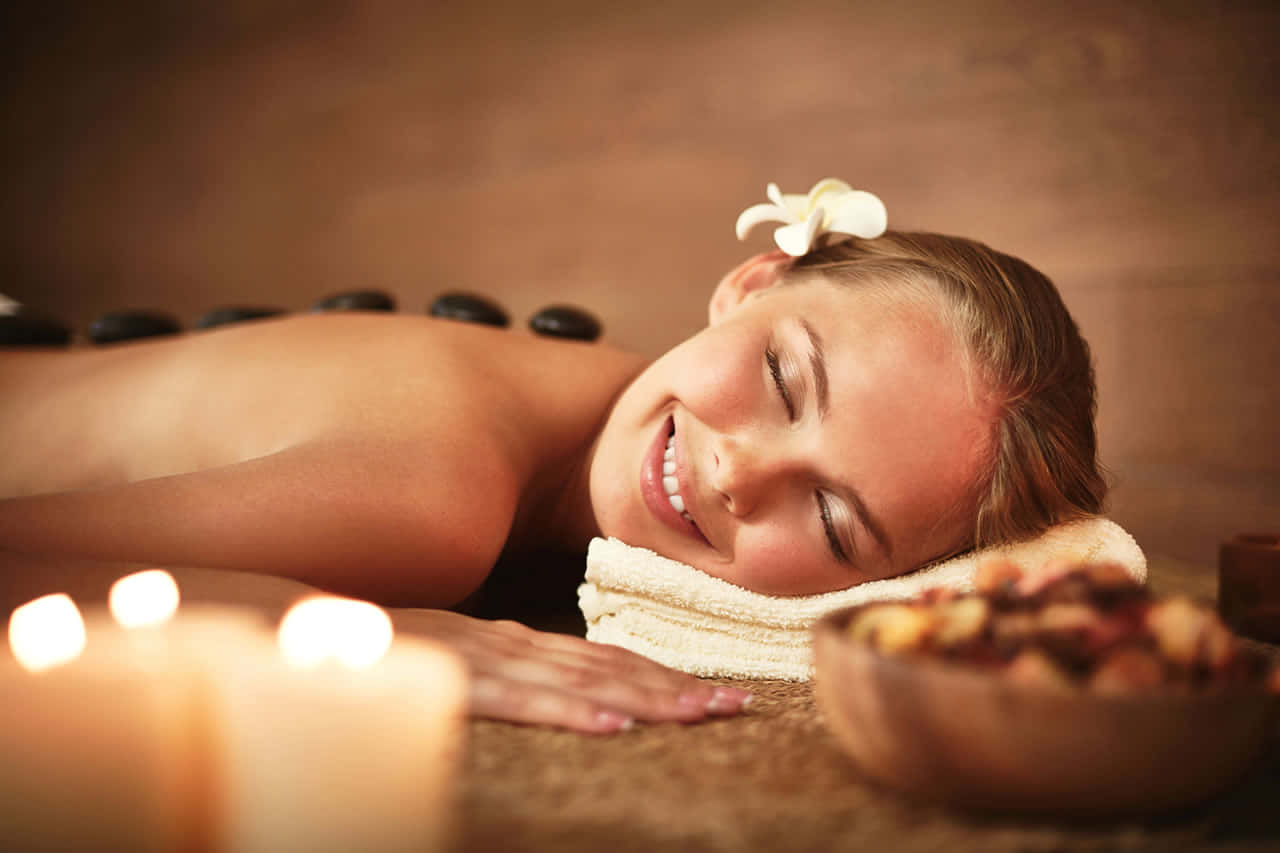 Massage 桌布 壁紙 — 一位寧靜的女性閉著眼睛微笑，溫暖的石頭沿著她的背部放置，在燭光水療環境中。