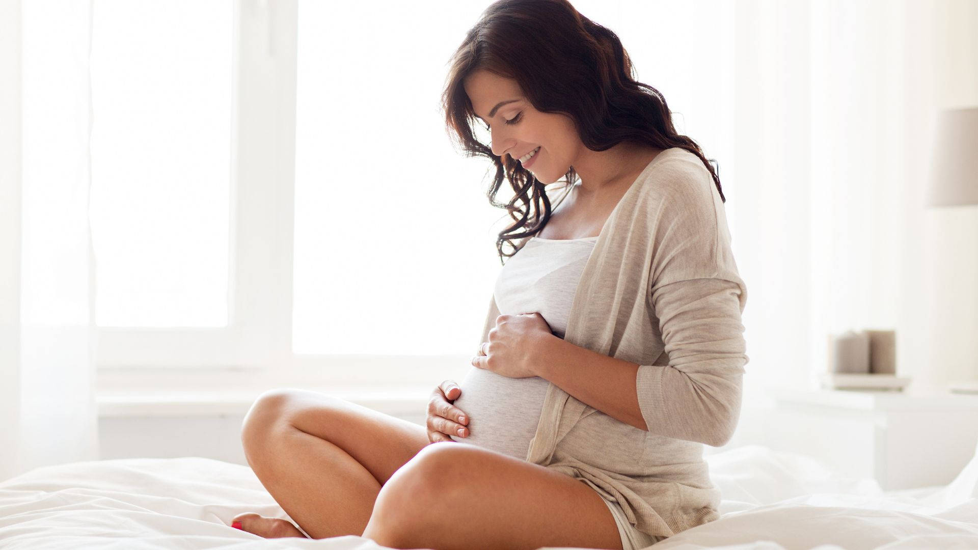 pregnancy 桌布 壁紙 — 一位寧靜的孕婦盤腿坐在床上，輕輕抱著腹部，沐浴在窗邊柔和自然光中。