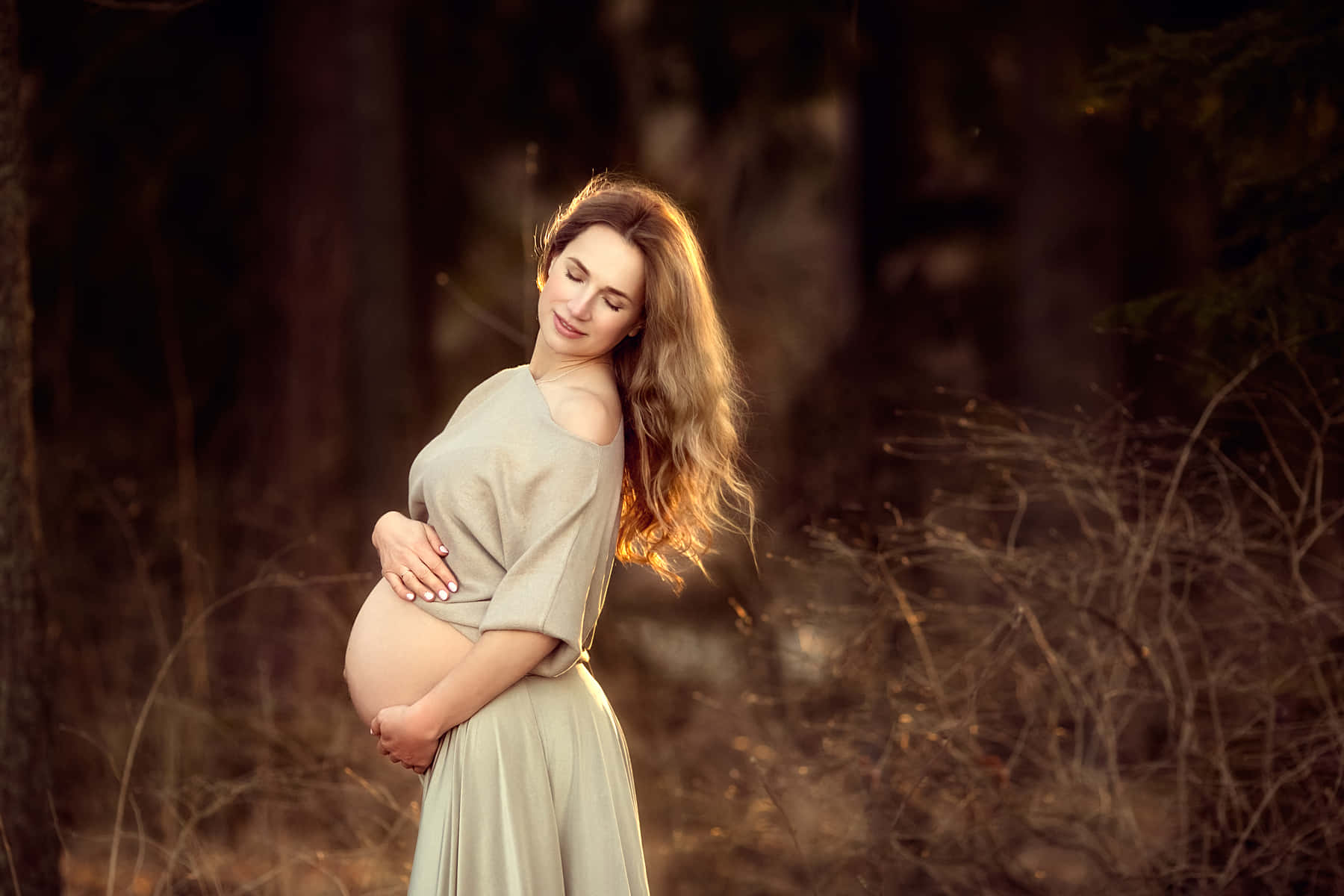 Pregnant 桌布 壁紙 — 一位寧靜的孕婦穿著飄逸的米色連衣裙，在陽光斑駁的森林中，金色光線穿過樹木，輕撫著她的腹部。