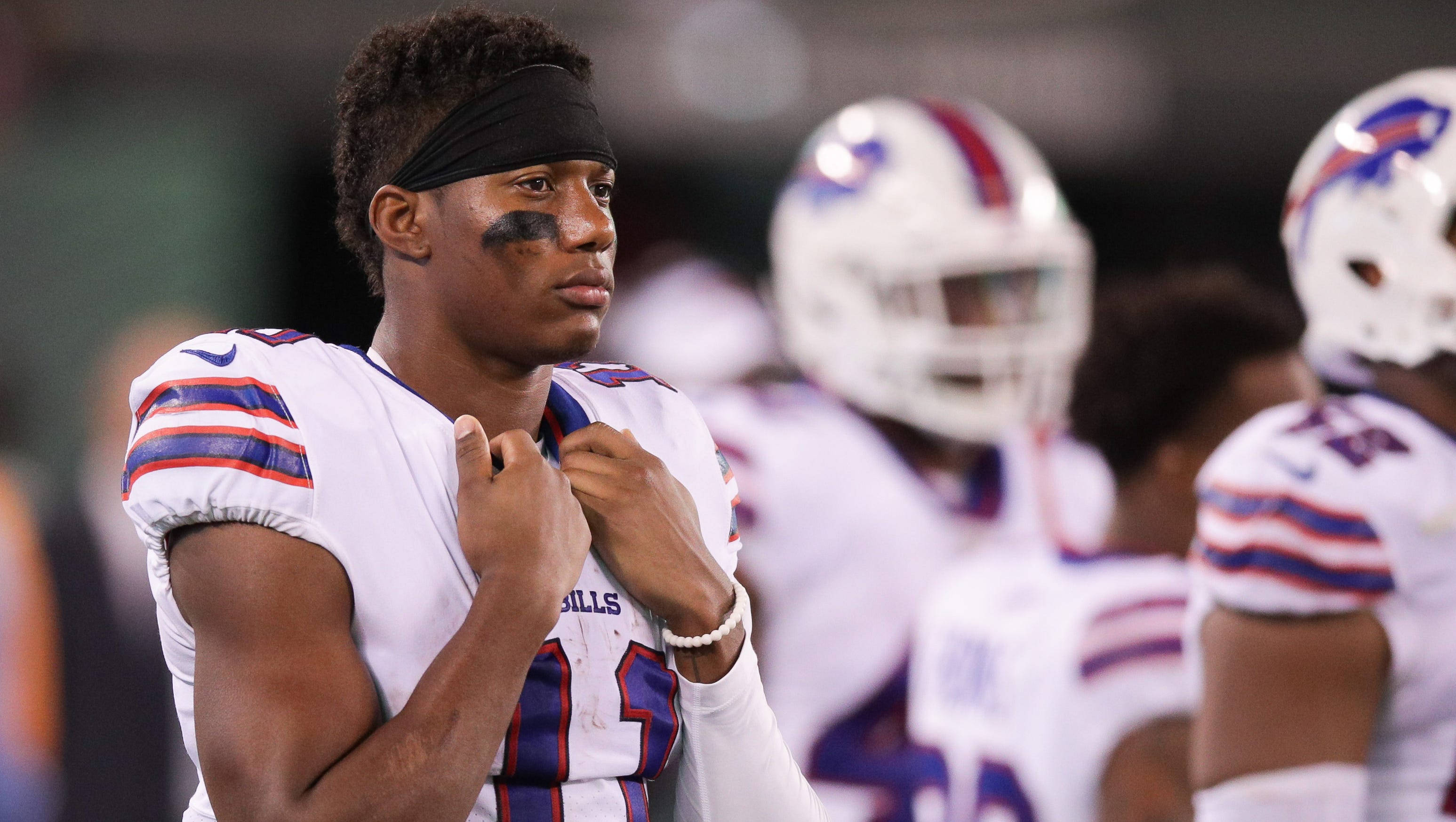 zay jones 桌布 壁紙 — 一位專注的Buffalo Bills美式足球員在球場燈光下調整球衣，背景有隊友模糊的身影。
