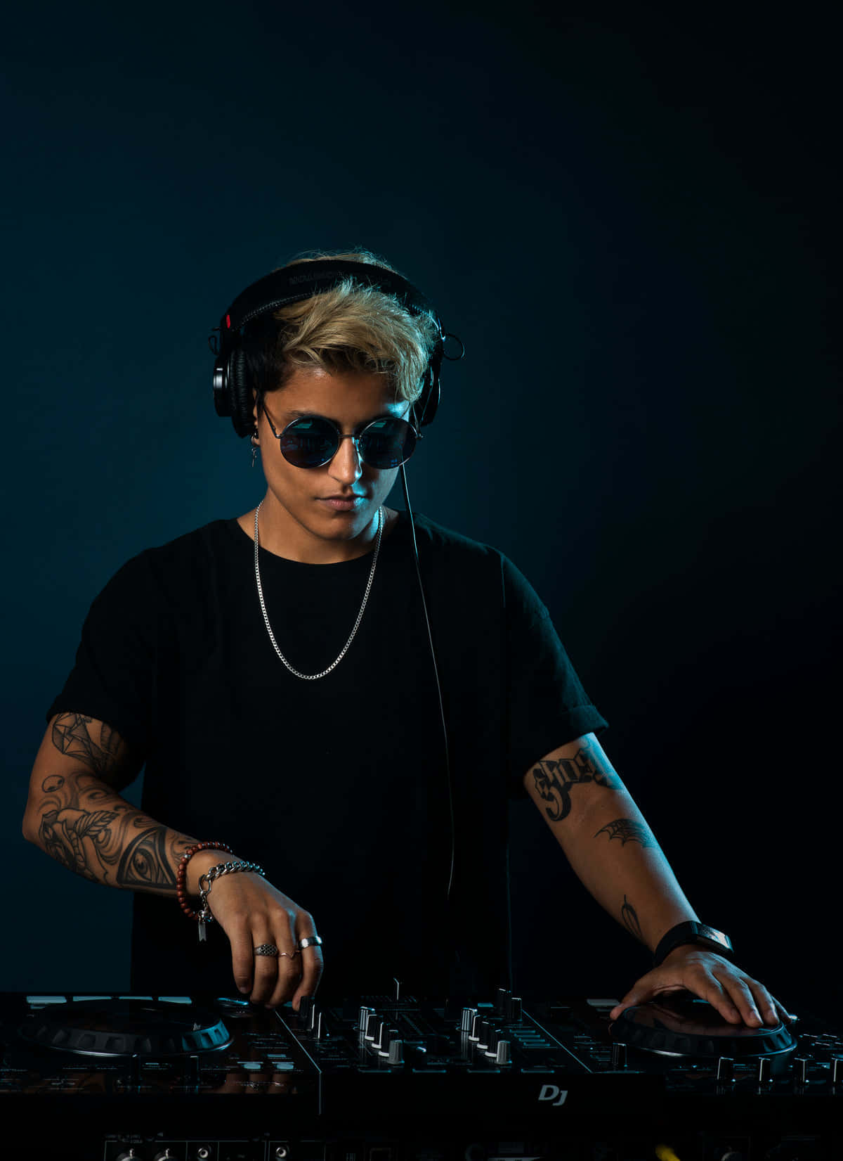 DJ 桌布 壁紙 — 一位專注的DJ，帶有刺青和太陽眼鏡，在戲劇性的藍色燈光下，在黑暗背景前操作唱盤。