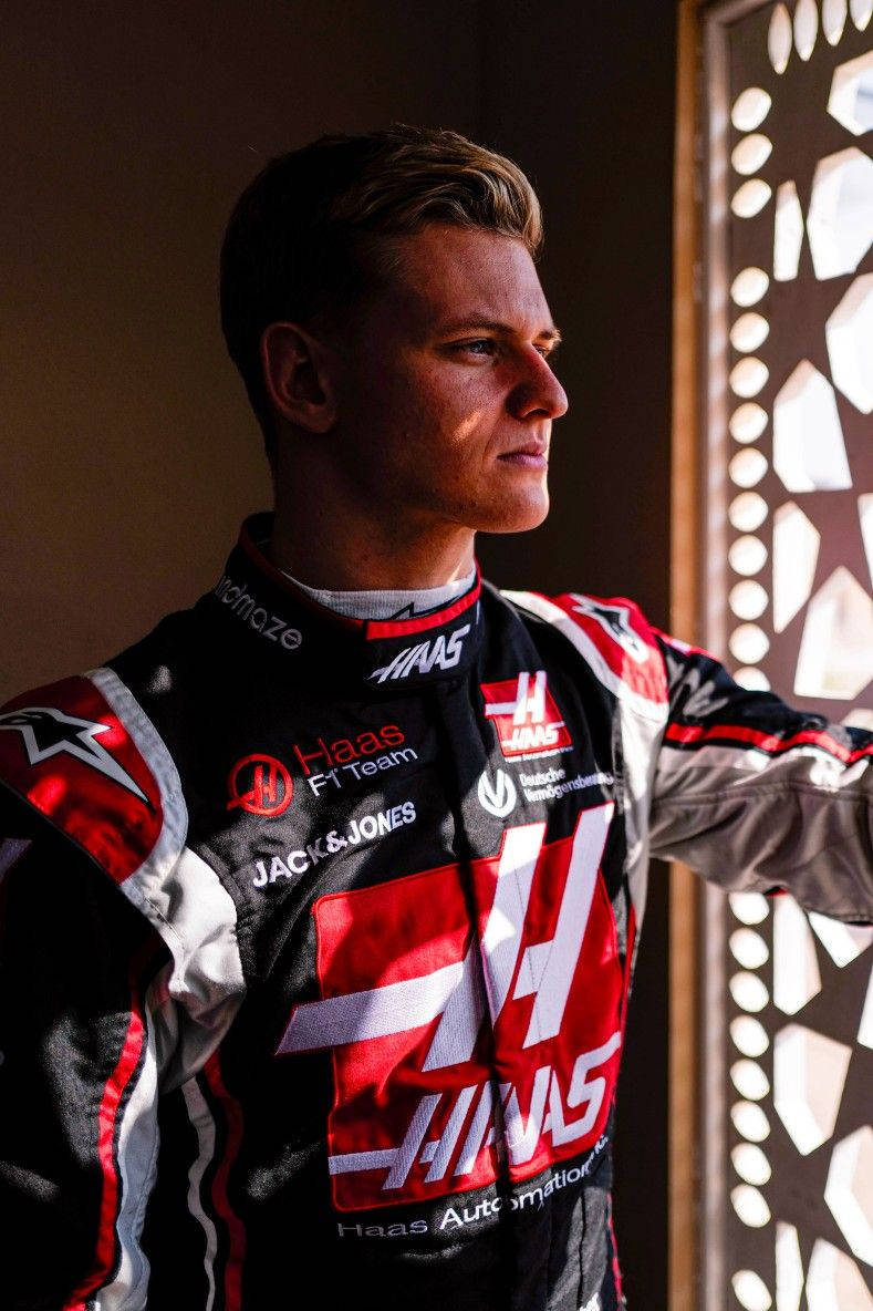 mick schumacher 桌布 壁紙 — 一位專注的F1車手身穿Haas賽車服，站在圖案窗戶旁，沐浴在溫暖的陽光中。