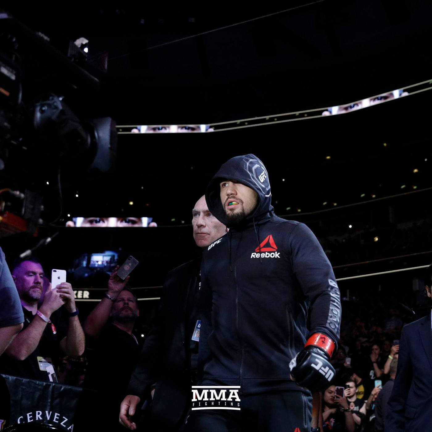 UFC 桌布 壁紙 — 一位專注的MMA拳手身穿黑色Reebok裝備和手套，在燈光昏暗的競技場中走過，周圍是攝影機和粉絲。
