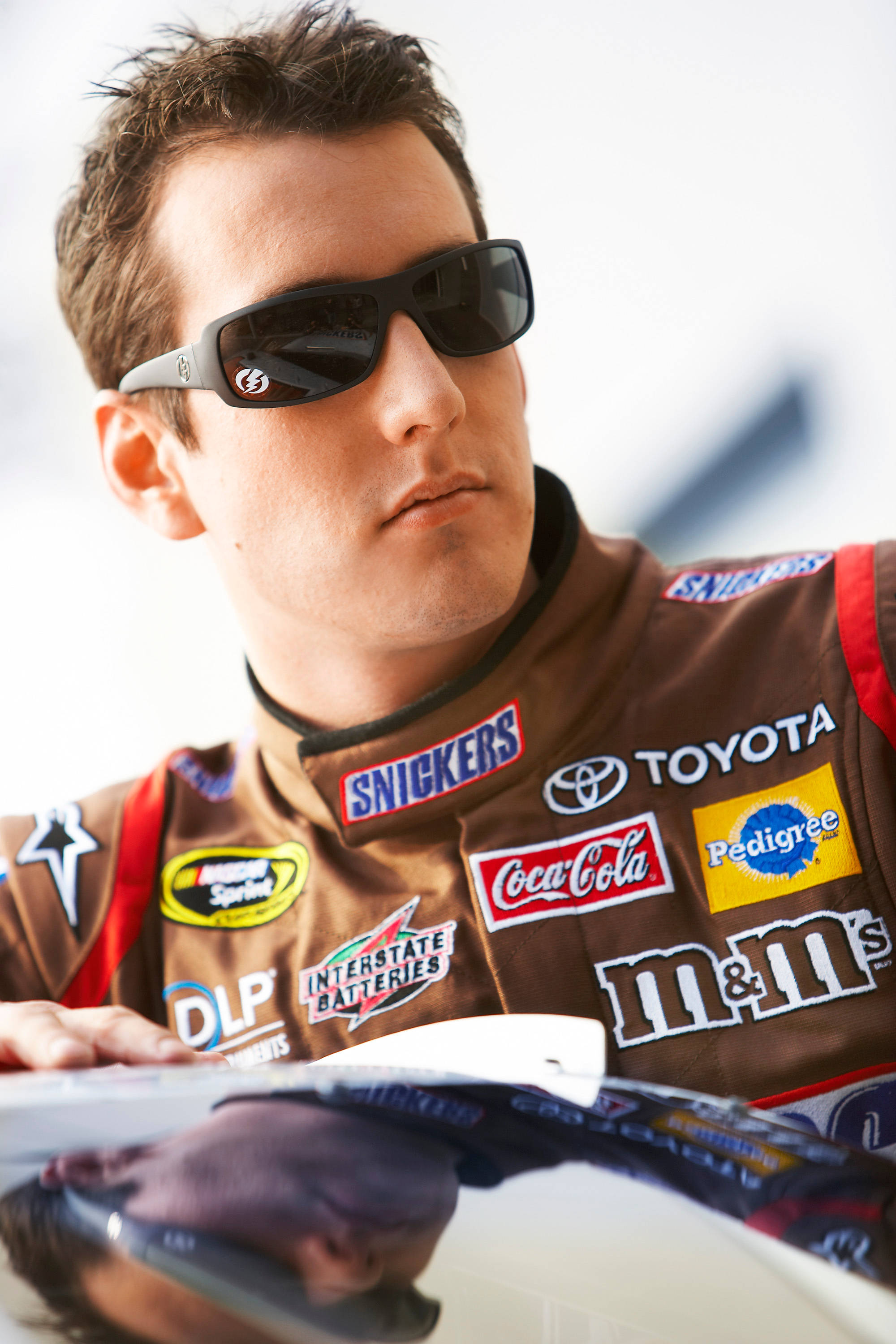 Racing 桌布 壁紙 — 一位專注的NASCAR車手穿著佈滿贊助商標誌的棕色賽車服和太陽眼鏡，俯身靠在汽車引擎蓋上。