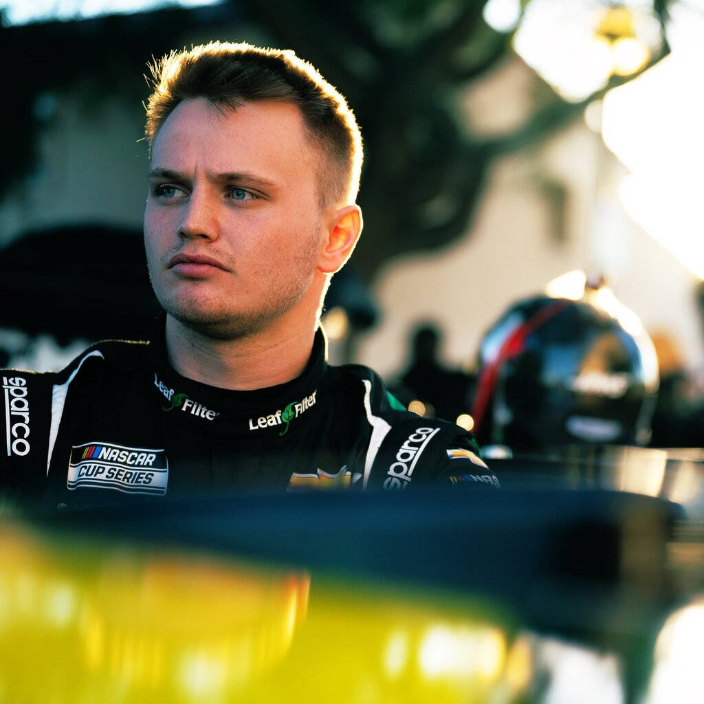 Racing 桌布 壁紙 — 一位專注的NASCAR車手穿著黑色賽車服，陽光照射下凸顯他的短髮和嚴肅表情，凝視著前方。