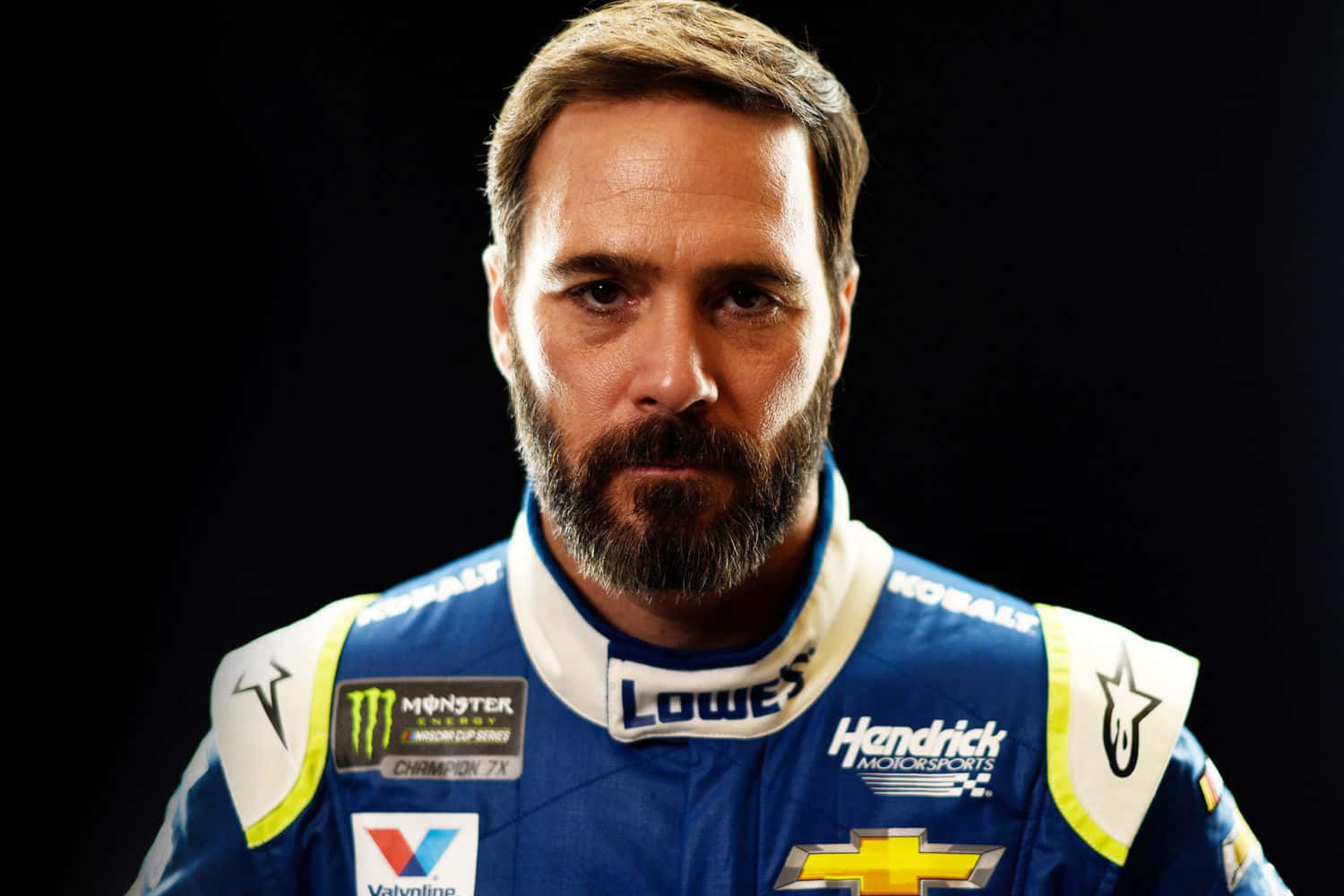 Jimmie Johnson 桌布 壁紙 — 一位專注的NASCAR車手身穿藍色賽車服，帶有贊助商標誌，在黑色背景下凝視著。