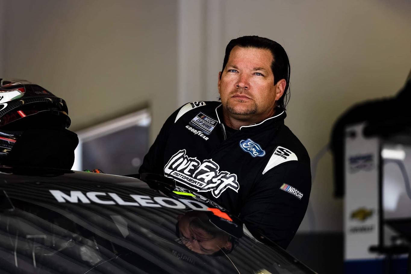 黑色 桌布 壁紙 — 一位專注的NASCAR車手身穿黑色「Live Fast」賽車服，在車庫裡靠著他的賽車。
