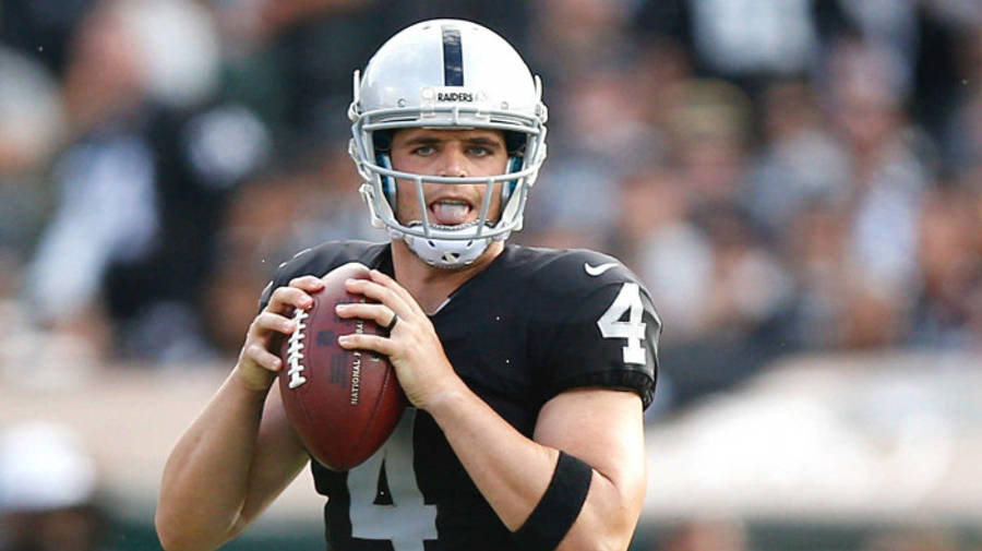 黑色 桌布 壁紙 — 一位專注的NFL四分衛穿著黑色Raiders球衣，在投球過程中以高度集中力抓住橄欖球。