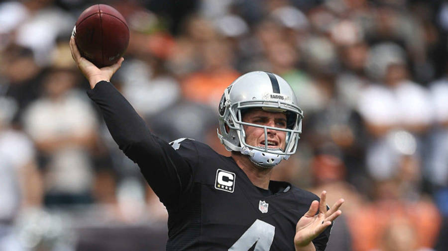 足球 桌布 壁紙 — 一位專注的NFL四分衛身穿黑色Raiders球衣，號碼4和隊長'C'，在白天比賽中擲出橄欖球，背景是模糊的體育場人群。