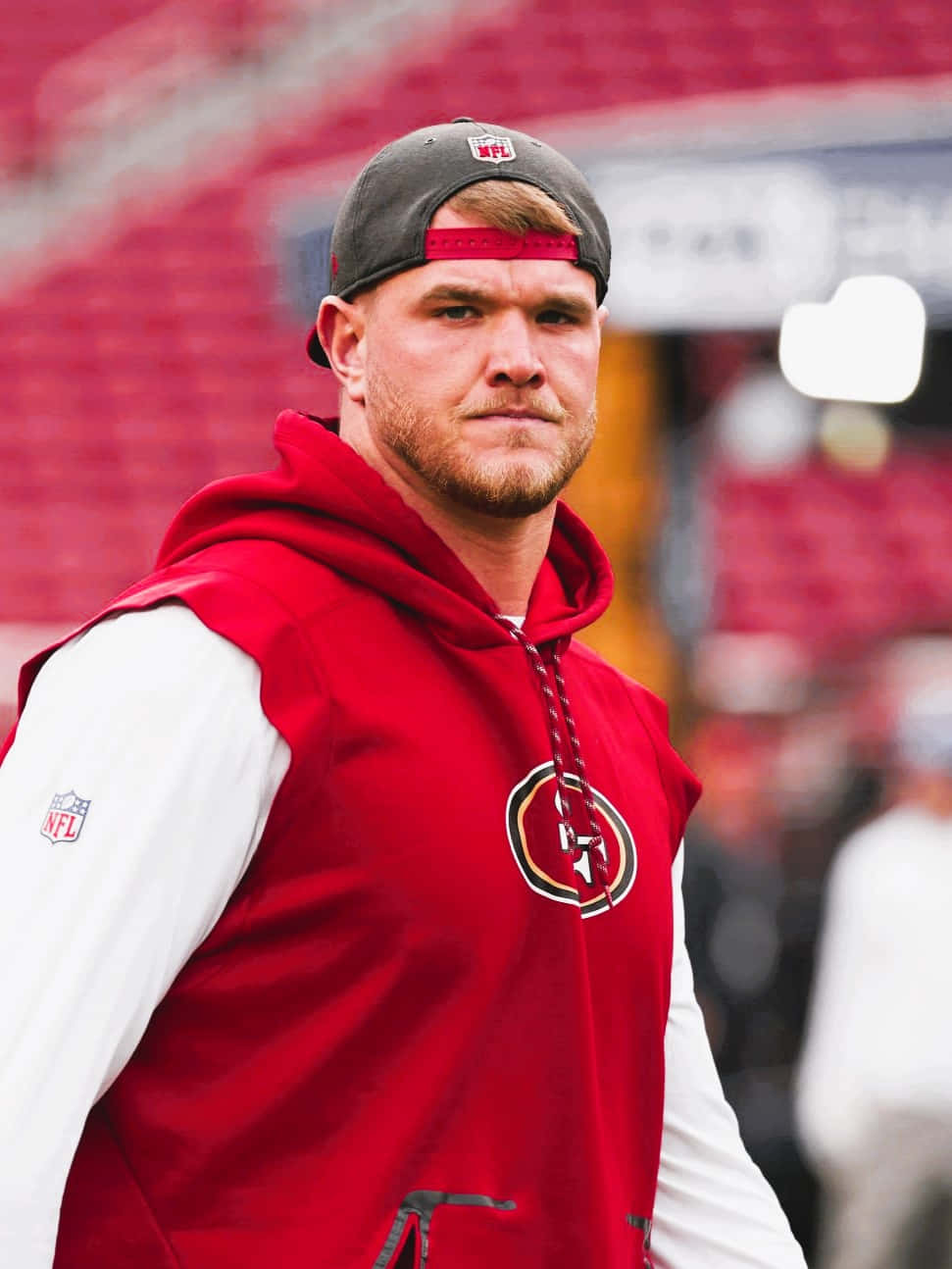Mike Mcglinchey 桌布 壁紙 — 一位專注的NFL球員身穿紅色舊金山49人連帽衫和帽子，站在球場上，背景是模糊的紅色座位。