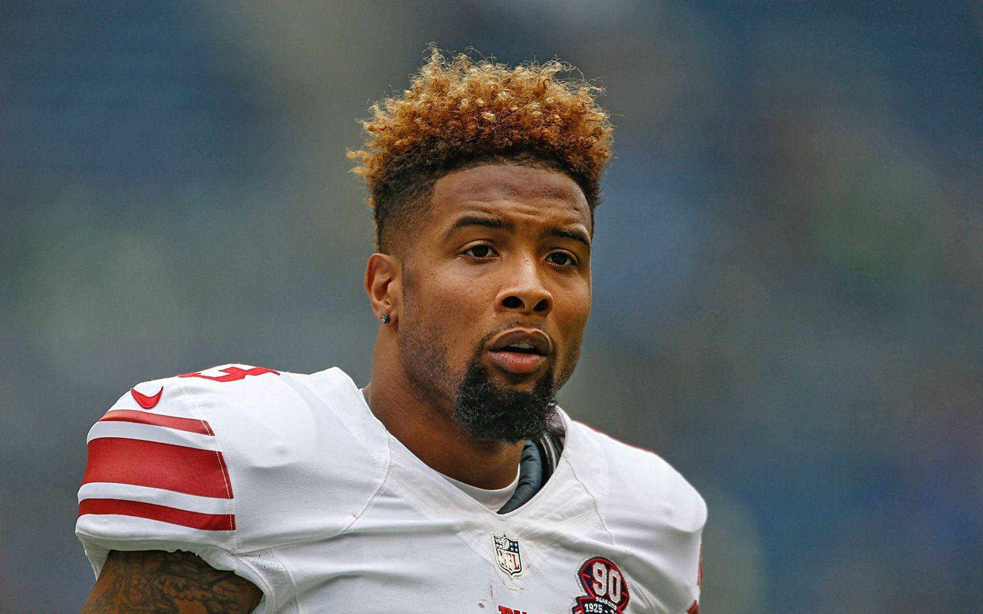 Odell 桌布 壁紙 — 一位專注的NFL美式足球員，金色捲髮和鬍鬚，在日間比賽中穿著帶有紅色條紋的白色球衣。