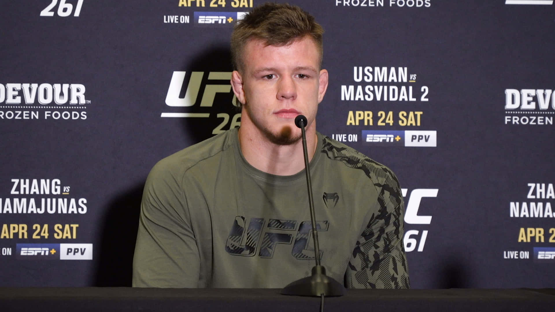 MMA 桌布 壁紙 — 一位專注的UFC拳手坐在記者會的桌子前，面前有麥克風，背景是品牌標誌。