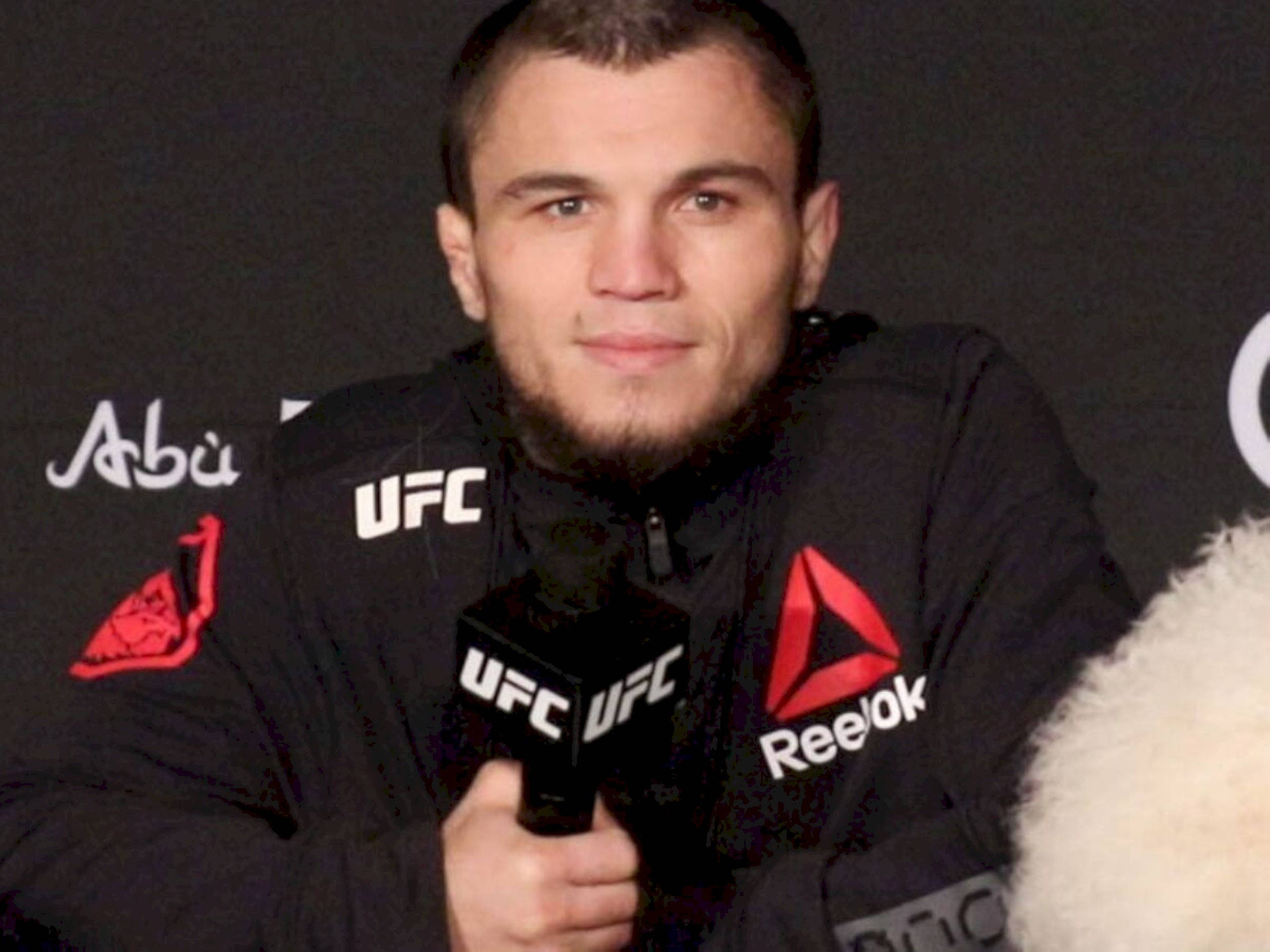 MMA 桌布 壁紙 — 一位專注的UFC拳手穿著黑色Reebok外套，在深色品牌背景前拿著麥克風。