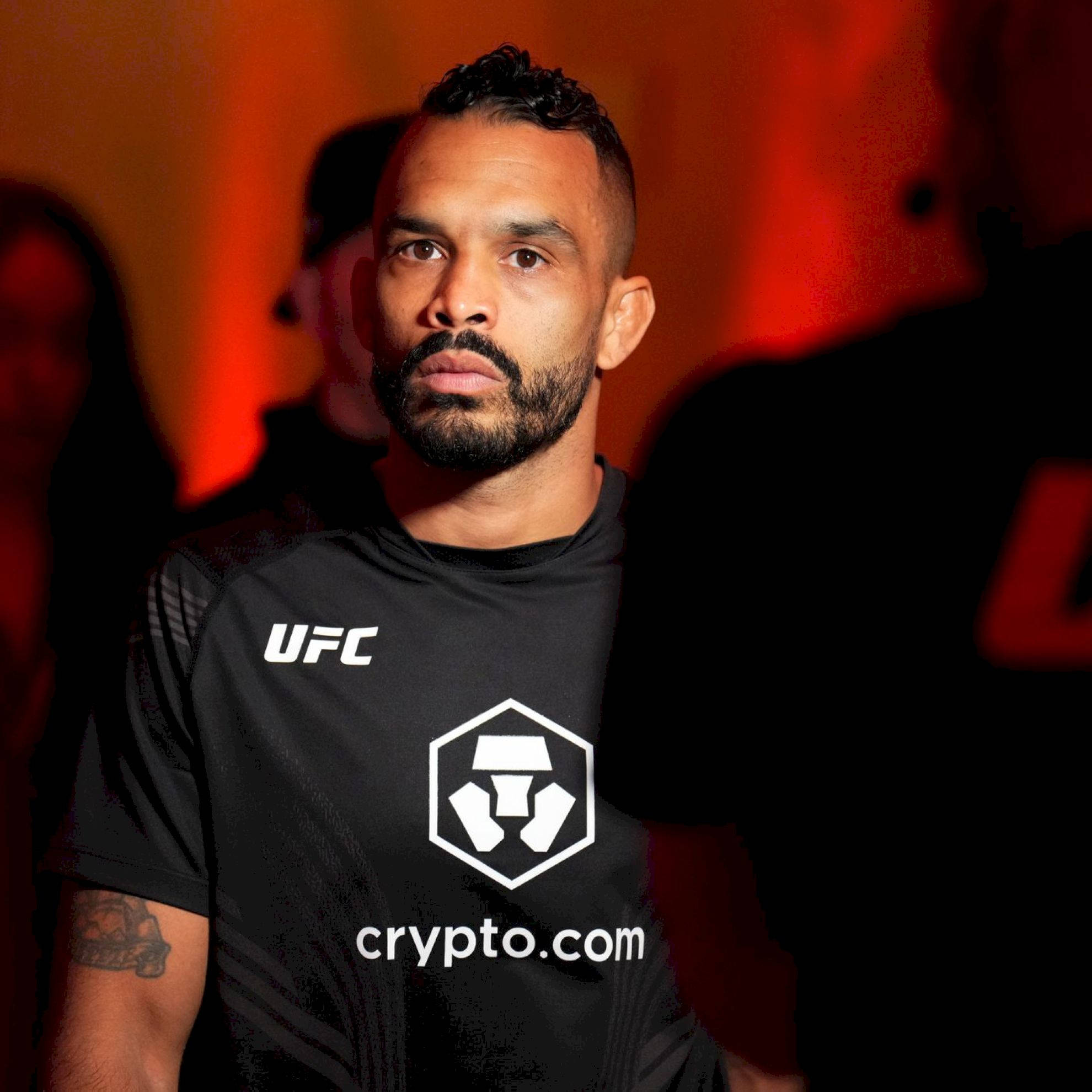 UFC 桌布 壁紙 — 一位專注的UFC拳手身穿黑色crypto.com品牌襯衫，站在溫暖的紅色燈光下，身後有模糊的人影。