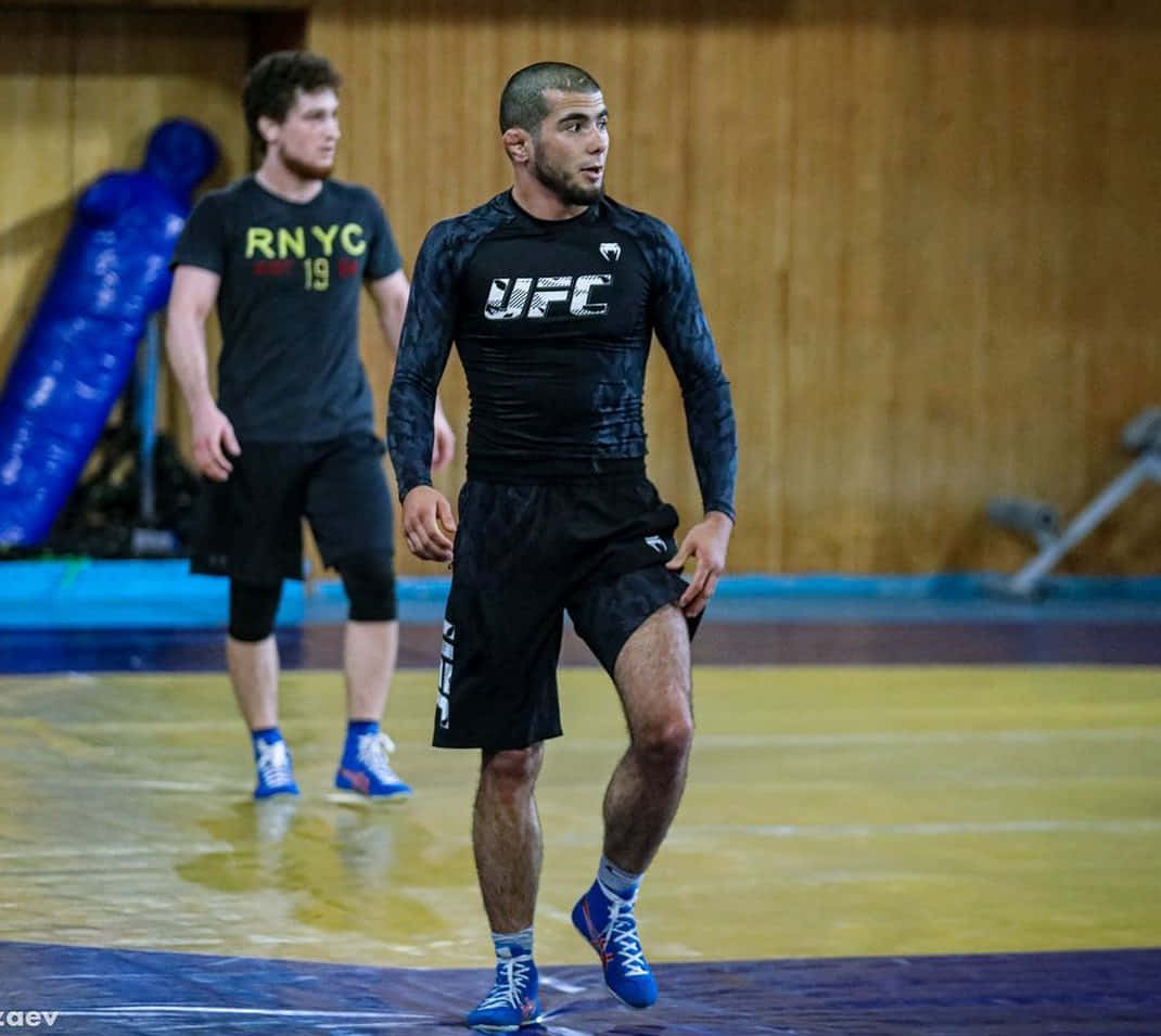 UFC 桌布 壁紙 — 一位專注的UFC拳手身穿黑色裝備，在健身房的摔角墊上大步走過，背景有一個藍色訓練假人。