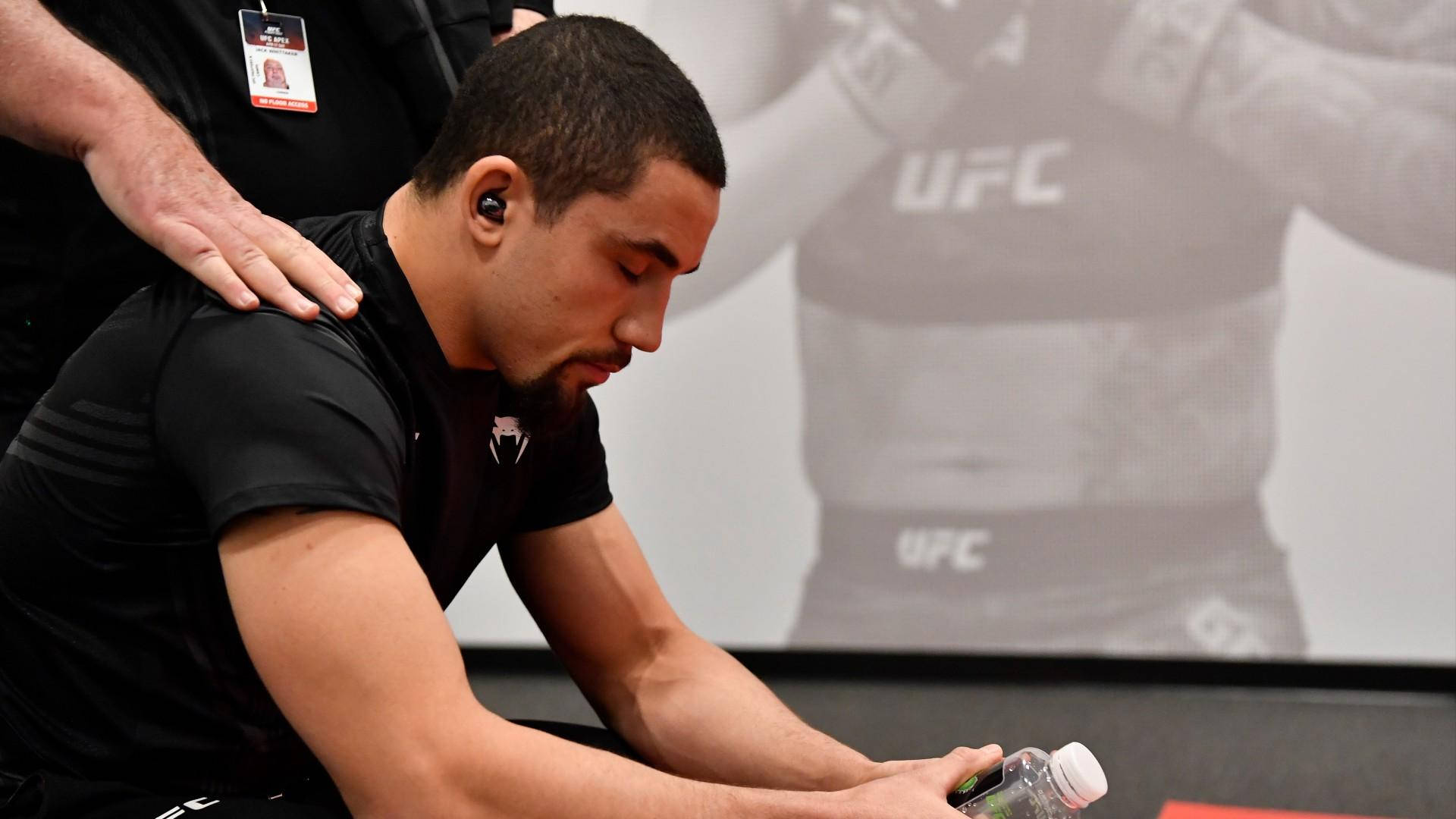 UFC 桌布 壁紙 — 一位專注的UFC拳手身穿黑色裝備，坐在墊子上，手持水壺，一手放在肩膀上，身處健身房環境。