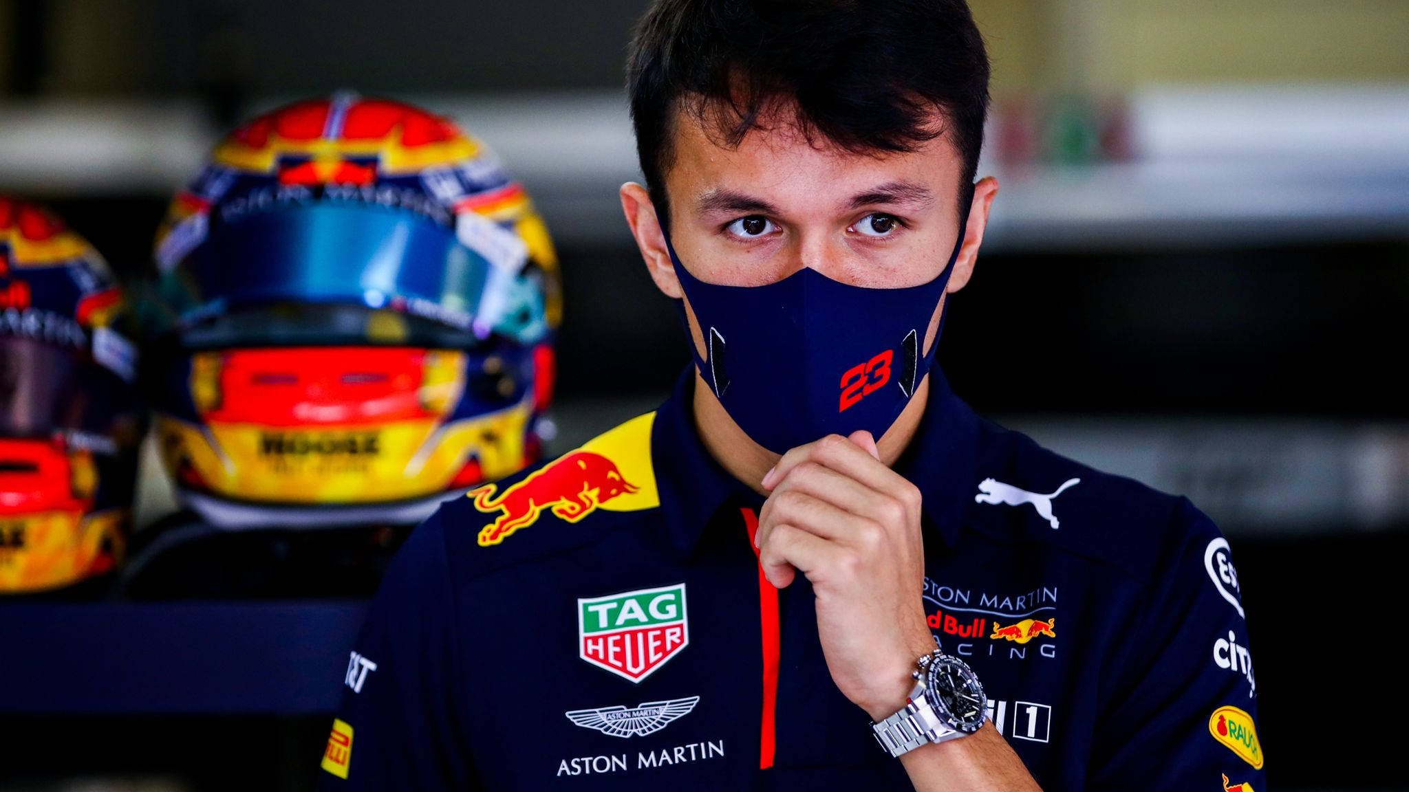 F1 桌布 壁紙 — 一位專注的一級方程式賽車手穿著紅牛車隊外套和口罩，背景有賽車頭盔。
