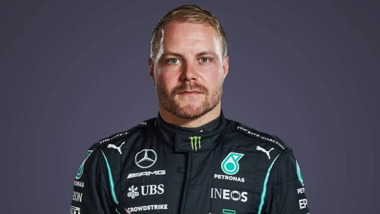 Valtteri Bottas 桌布 壁紙 — 一位專注的一級方程式賽車手身穿黑色賽車服，帶有青綠色點綴和贊助商標誌，在純深灰色背景前擺姿勢。