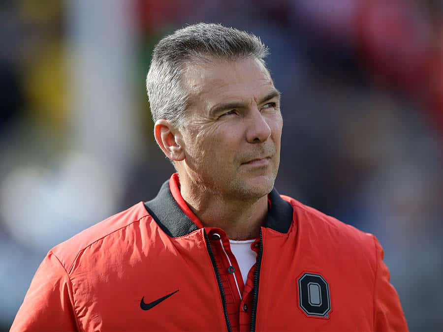 Urban Meyer 桌布 壁紙 — 一位專注的中年男子穿著紅色俄亥俄州立大學外套，在明亮的日光下凝視著運動場。