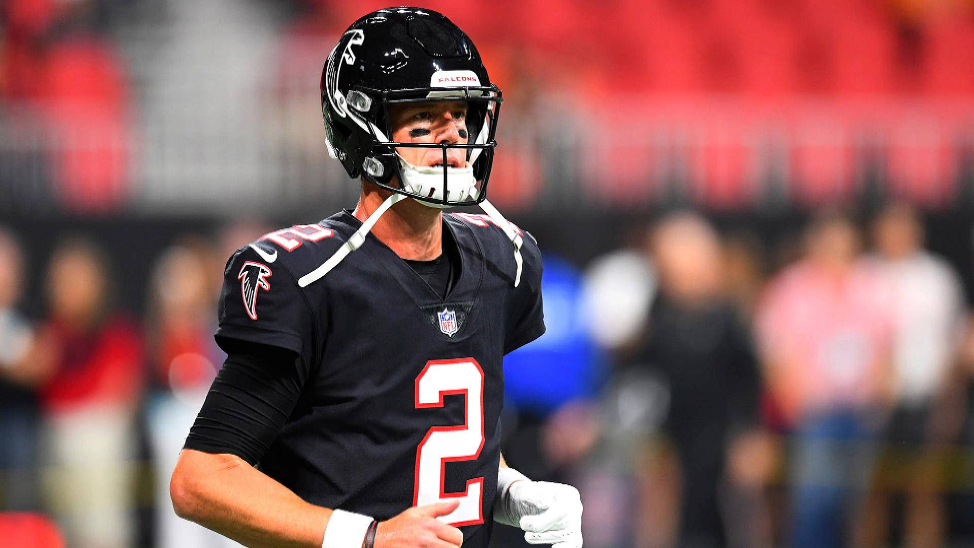 Matt Ryan 桌布 壁紙 — 一位專注的亞特蘭大獵鷹四分衛身穿黑色二號球衣，站在燈火通明的足球場上，身後是模糊的體育場人群。