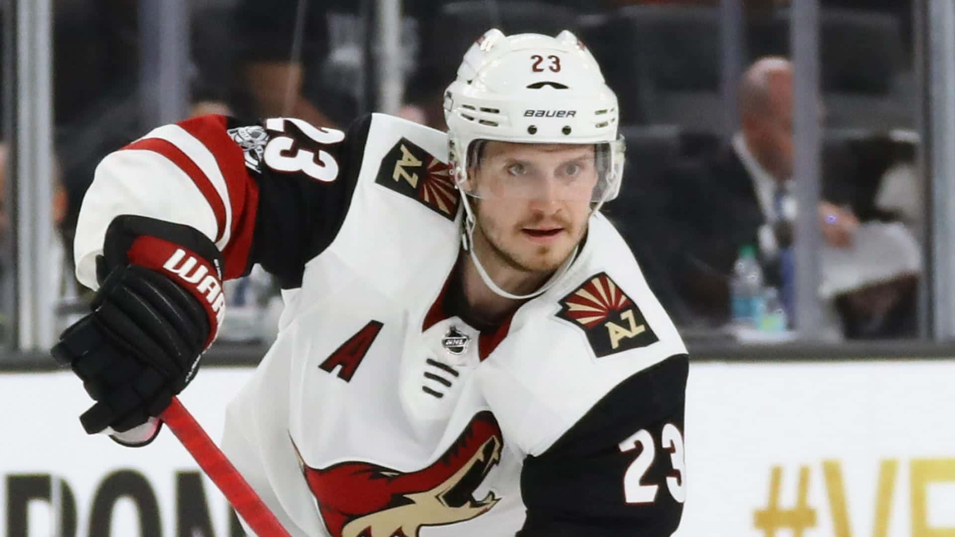 hockey 桌布 壁紙 — 一位專注的冰球運動員身穿白色和紅色的亞利桑那郊狼隊23號球衣，在激烈的比賽中向前傾身緊握球桿。