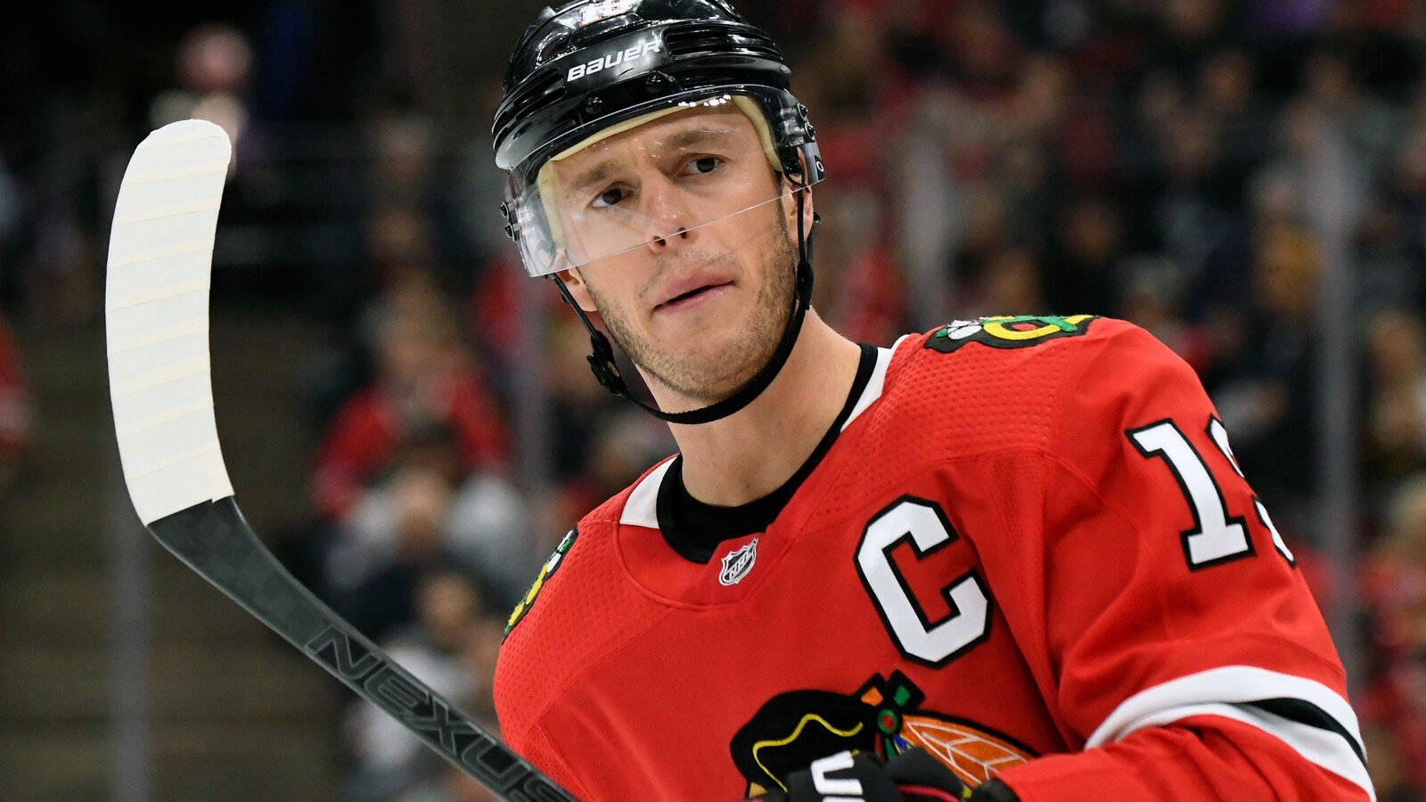 hockey 桌布 壁紙 — 一位專注的冰球隊長穿著紅色芝加哥黑鷹隊球衣，在明亮的場館燈光下於激烈比賽中握著球桿。