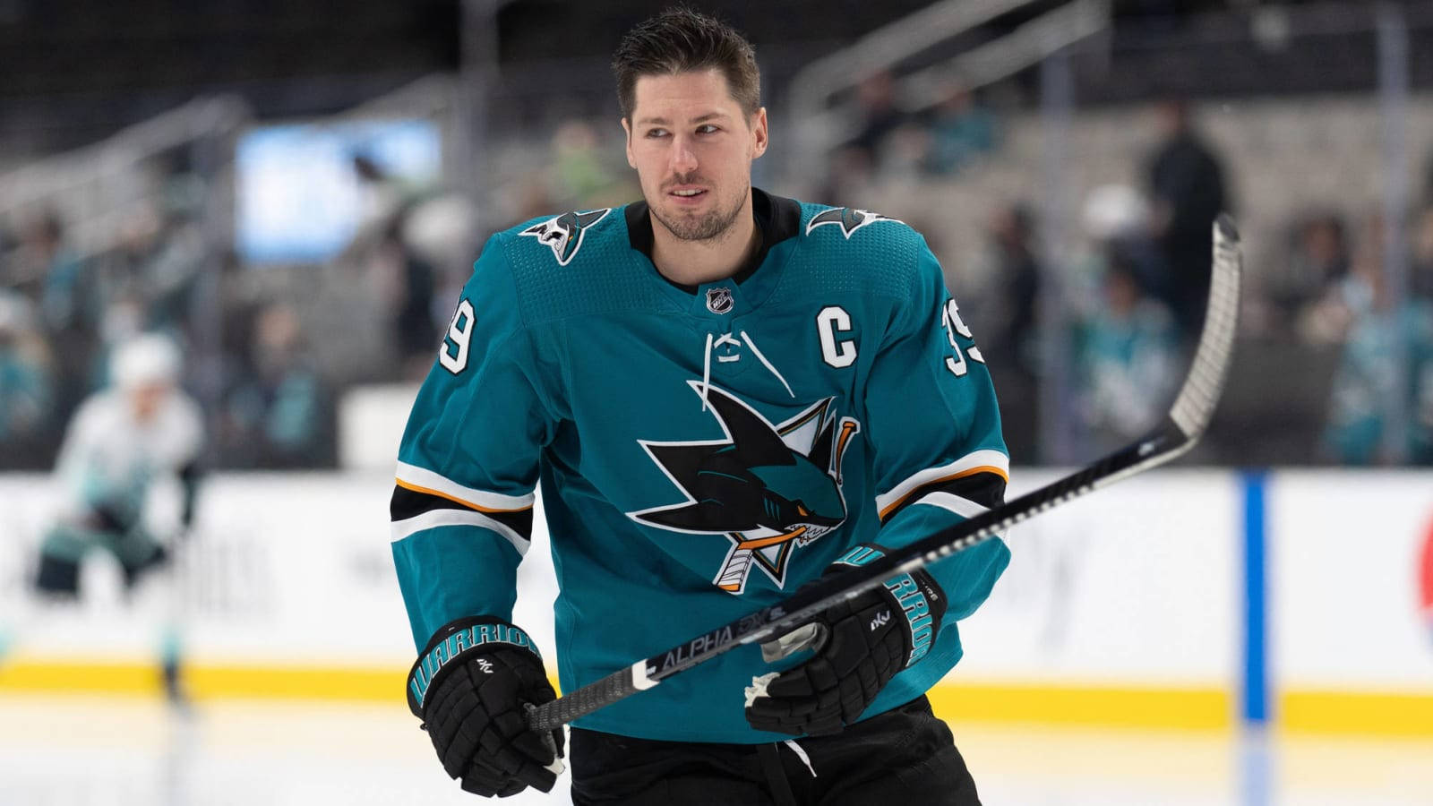 hockey 桌布 壁紙 — 一位專注的冰球隊長穿著青色鯊魚隊球衣，在比賽中於冰上握著球桿，背景是模糊的觀眾。